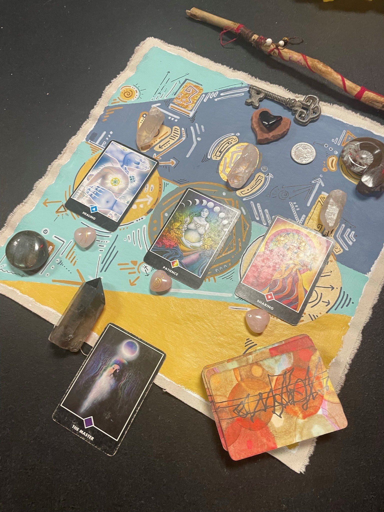 A tarot or oracle card reading setup on a colorful cloth or mat with various cards displayed, crystals, stones, a key, and other small objects, along with a decorated stick or wand.