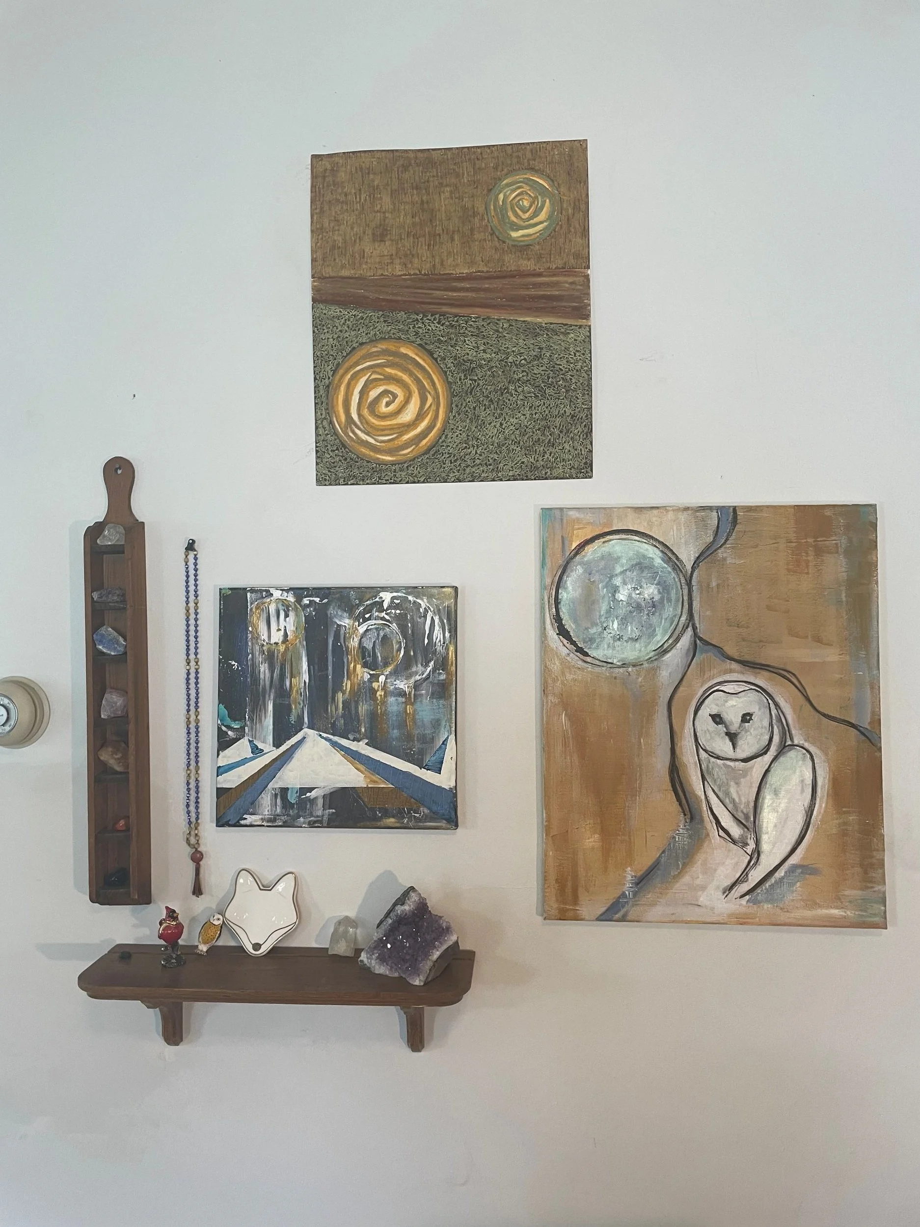 Gallery wall with abstract paintings, small decorative items, a wooden shelf with crystals and figurines, and a mirror on a white wall.
