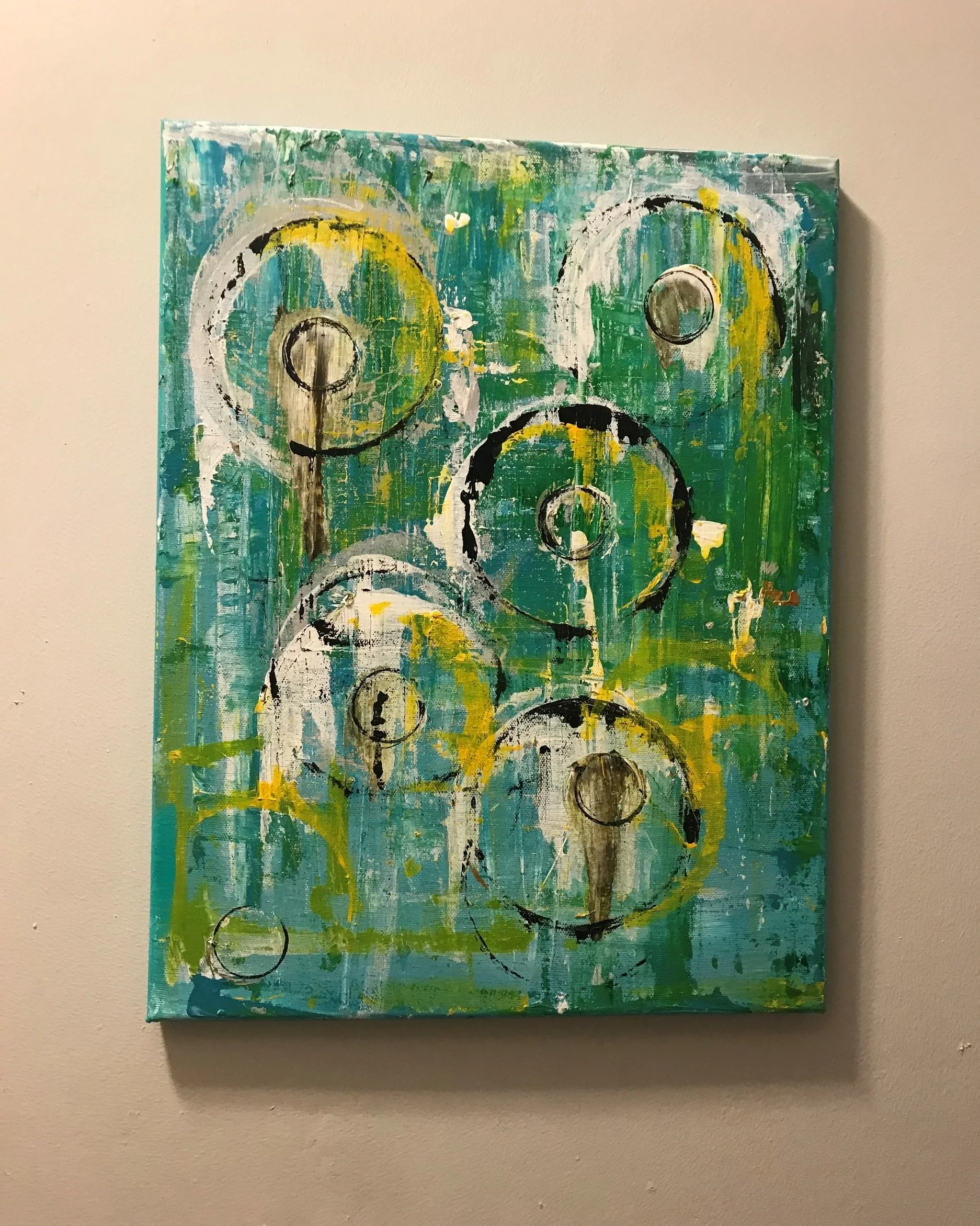 Abstract painting with circular shapes, green, yellow, white, and black colors, and textured brushstrokes, hanging on a beige wall.