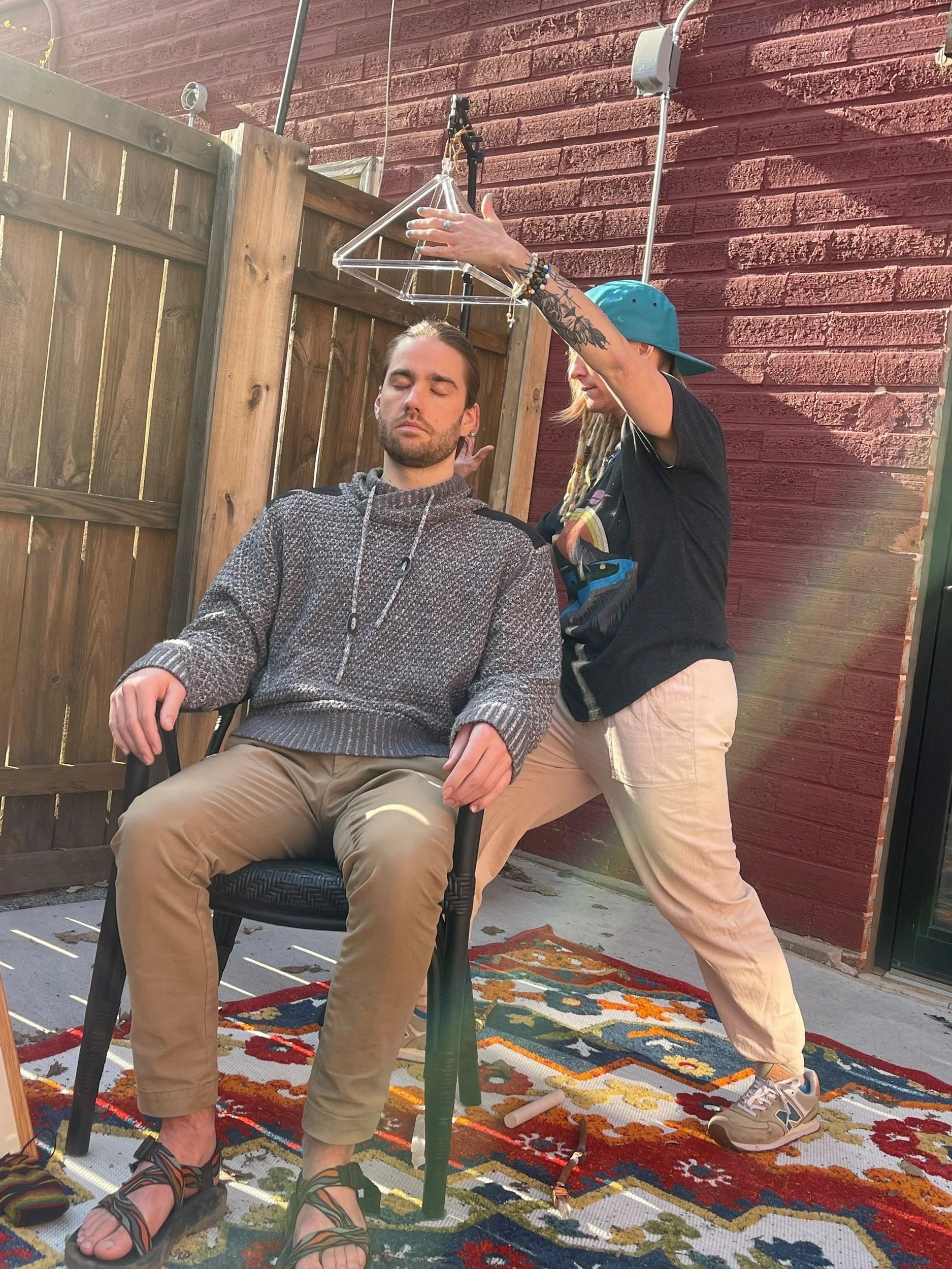 A man with closed eyes sitting in a chair with a patterned rug beneath him, while a woman with a blue cap and tattoos adjusts a triangular hanging object above him in an outdoor setting with a wooden fence and a red brick wall.