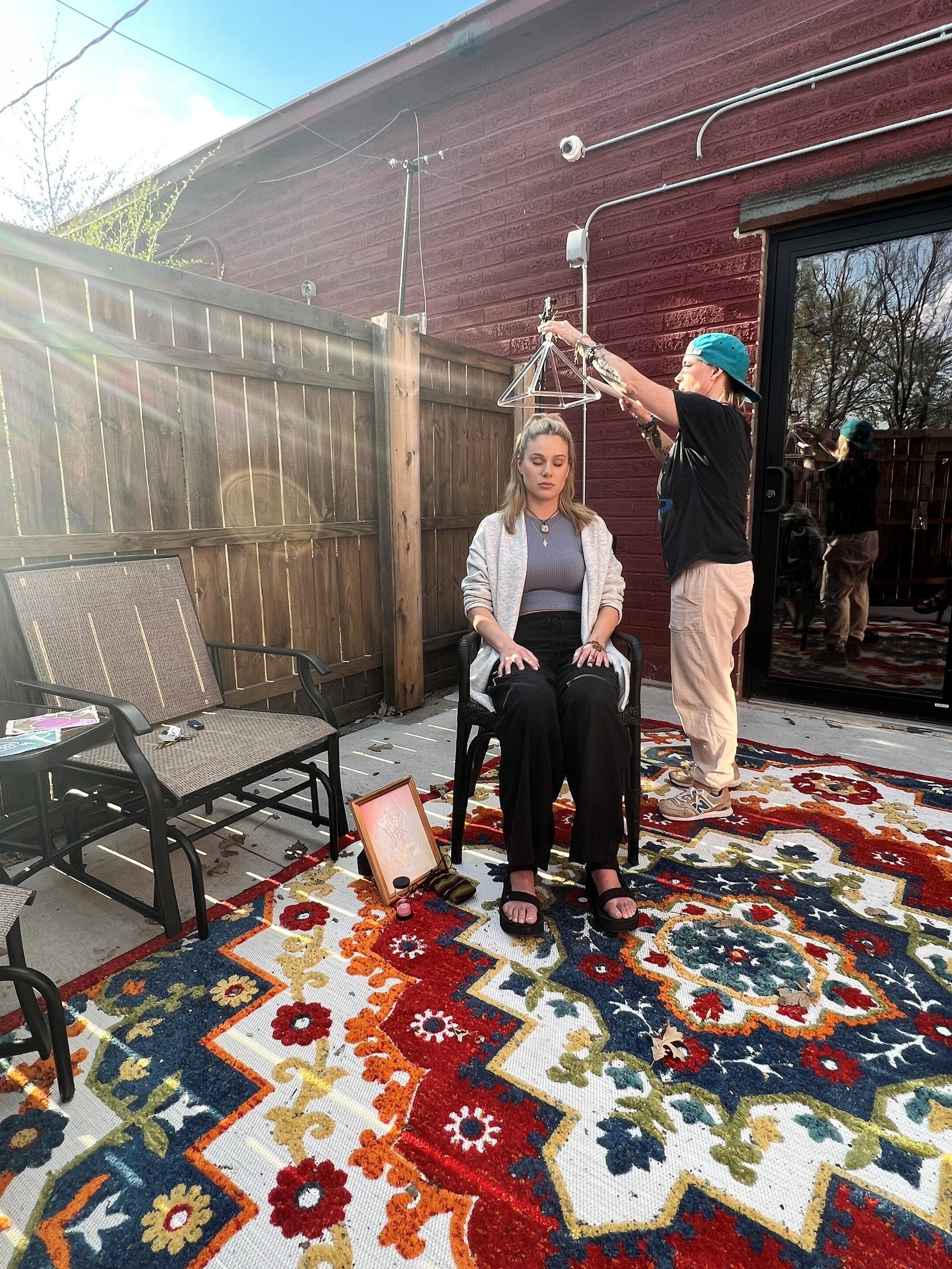 A woman with blonde hair sitting on a chair with her eyes closed outdoors, while a person with a teal cap and tattoos hangs a triangular metal clothing rack above her head. The scene is next to a red wooden house with a sliding glass door, decorated 