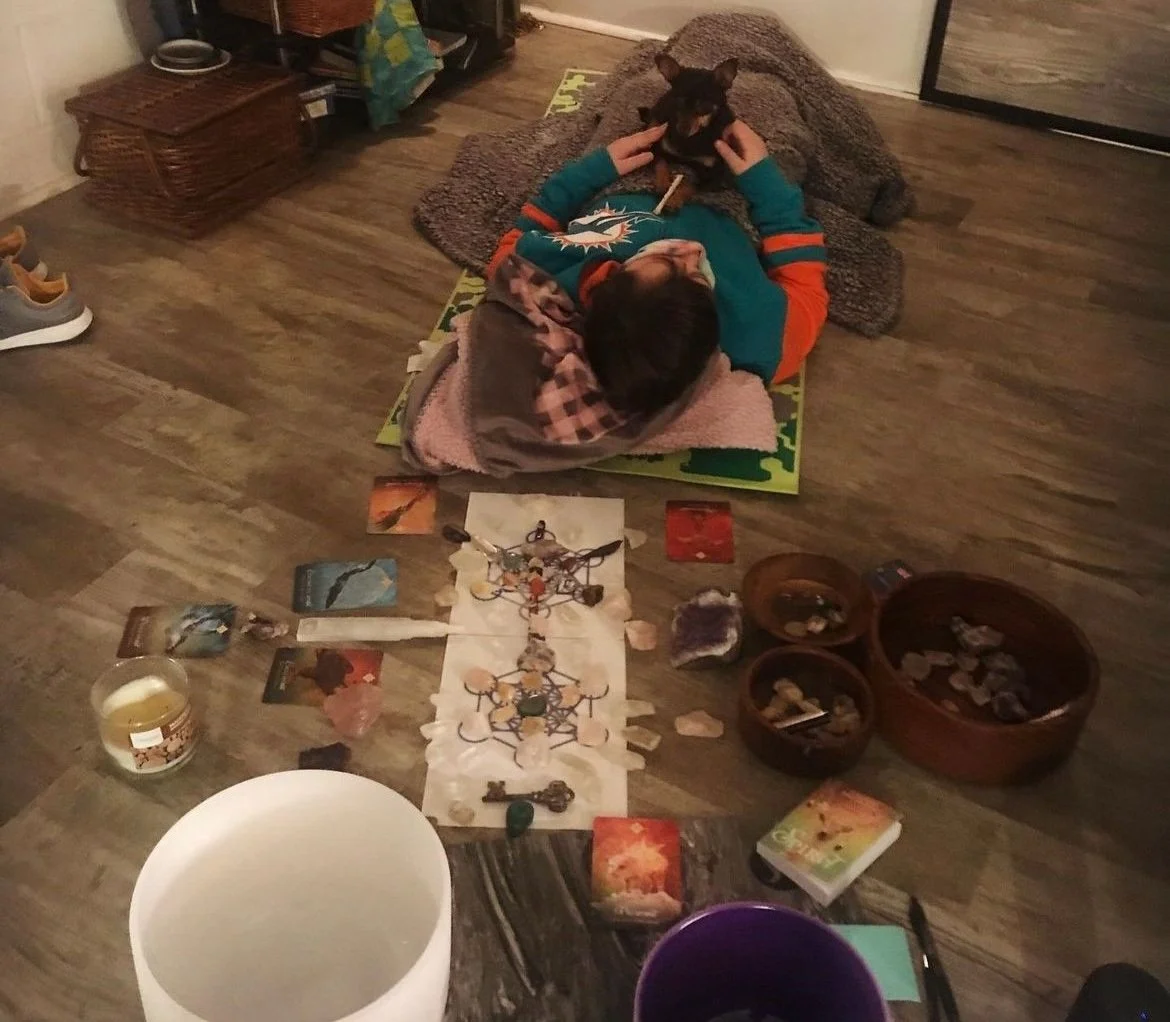 Person lying on their back on the floor with a blanket, holding a small black dog above their head, surrounded by tarot cards, crystals, and tarot-like booklets, with bowls of stones nearby.