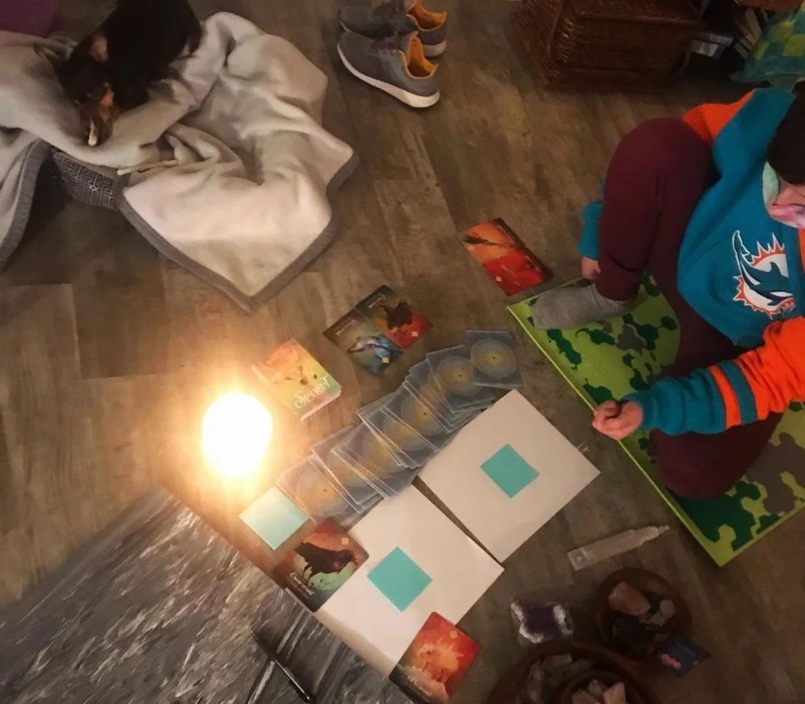 A person playing a card game on a green playmat, with cards, game pieces, and character cards spread on the floor. A sleeping cat rests on a blanket nearby, and a lit candle illuminates the scene.