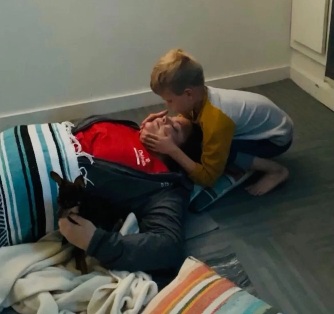 A boy is leaning over a girl who is lying on the floor, holding her face with one hand and touching her forehead with the other, possibly checking her temperature. There are blankets and a blanket on the floor, and a small dog is sitting near them, b
