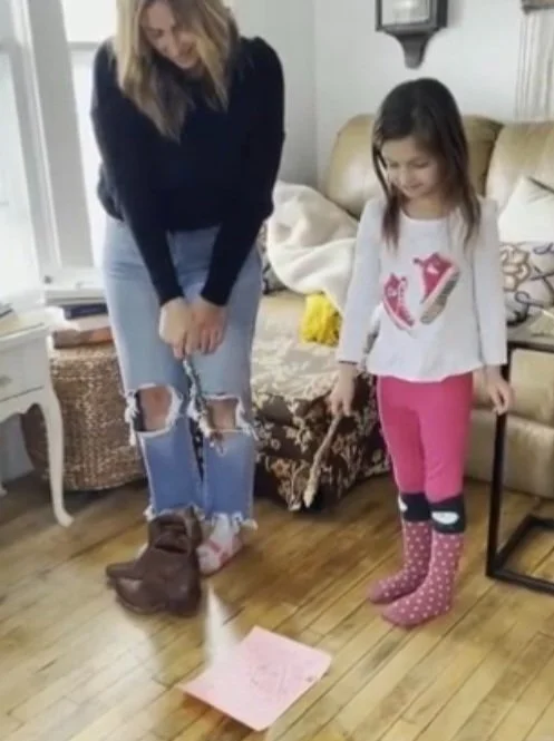 A woman and a young girl are playing a game in a living room. The woman is standing, wearing ripped jeans and socks, with her boots on the floor. The girl, dressed in a white shirt with pink leggings and pink polka-dot socks, is holding a stick with 