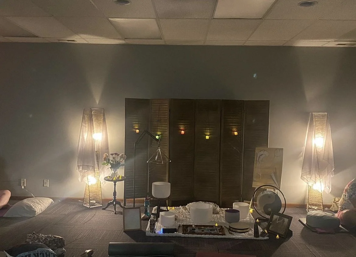 Group Sound Bath Room. A dimly lit room with two tall floor lamps on either side of a wooden partition. There are various objects on the floor, including bowls, a bottle, a picture frame, and a chair. The room has a relaxed, cozy atmosphere.