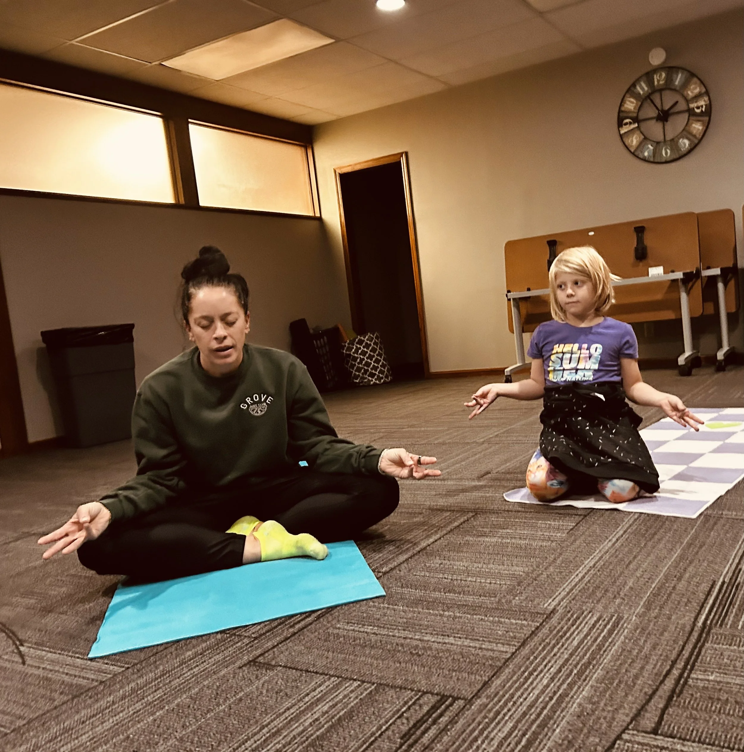 A woman and a young girl are practicing yoga on colorful mats in a room with beige walls, a large clock, and some furniture in the background. The woman is in a seated meditation pose with her eyes closed, wearing a dark green sweatshirt, and the gir