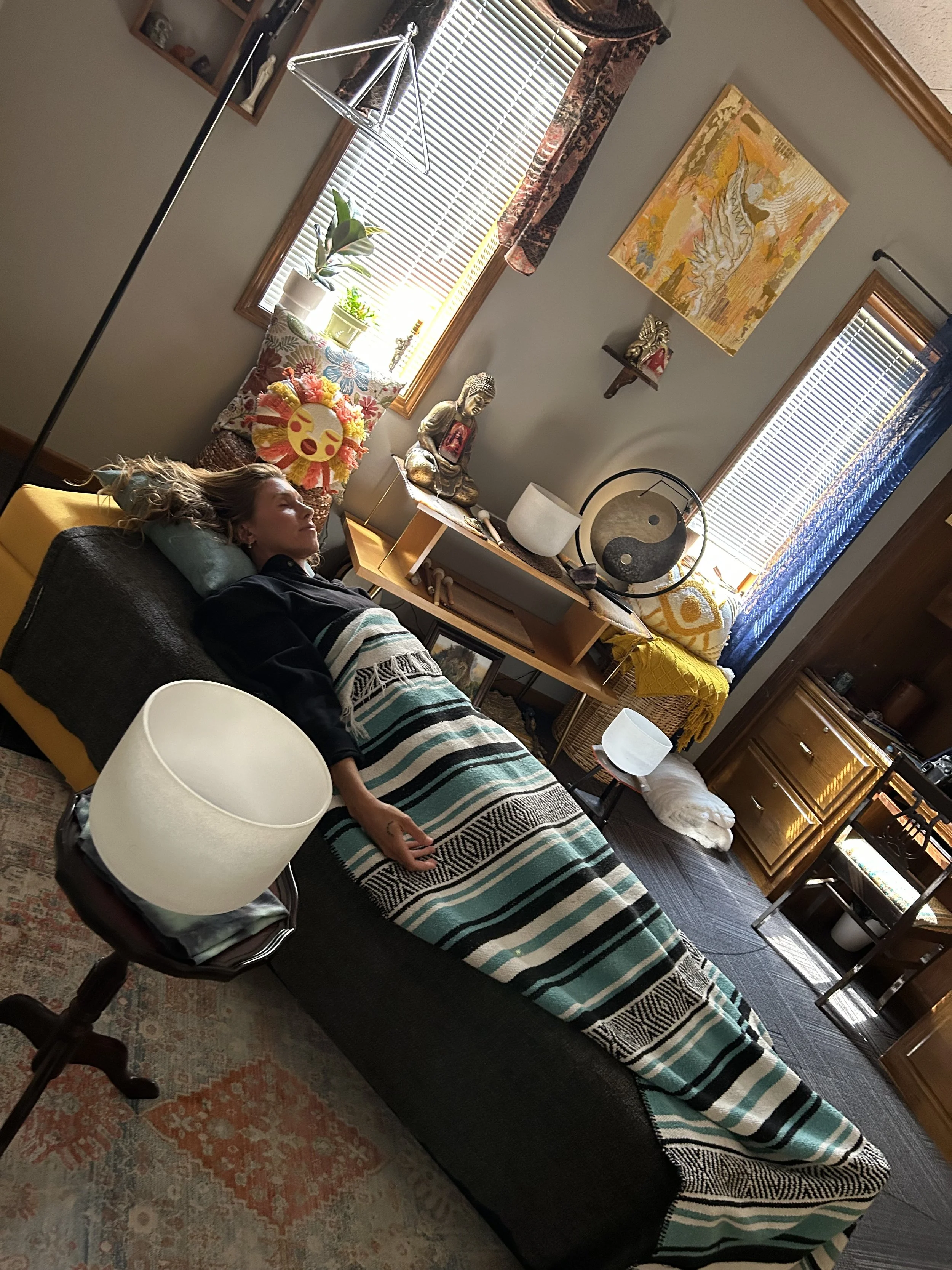 Crystal Bowl Healing. A woman sleeping on a couch covered with a striped blanket in a living room with various decorations, artwork, plants, and furniture.