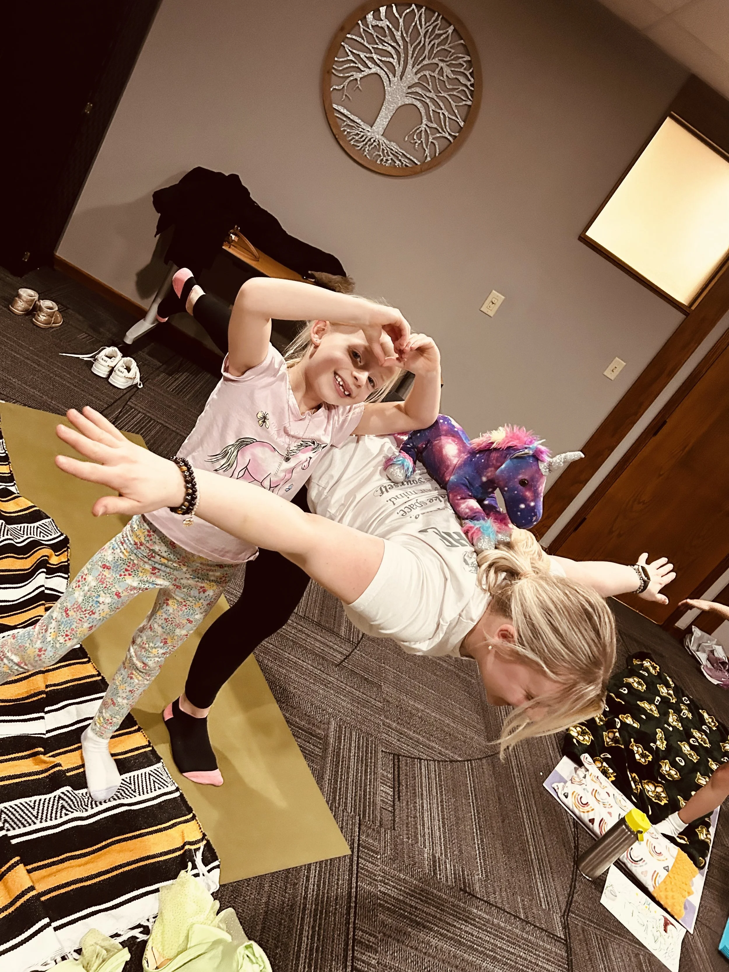 A young girl with blonde hair smiling and making a peace sign with her hand, holding a colorful unicorn plush toy and standing on a yellow mat in a room with dark walls and a framed tree artwork. An adult woman is bending over a table, and other chil