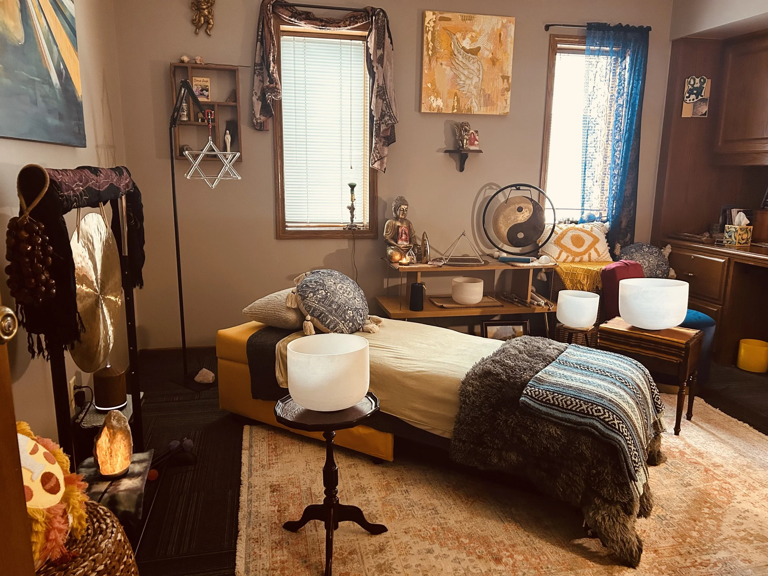 A cozy and eclectic living room with a mix of global decor. It features two windows with curtains, a variety of art on the walls, a table with spiritual and decorative objects including a Buddha statue, a yin-yang decor piece, and lamps. There’s a beige couch with patterned pillows, a colorful blanket, and a yellow ottoman. The room has a wooden floor, a patterned rug, and a tall speaker or lamp near the door.
