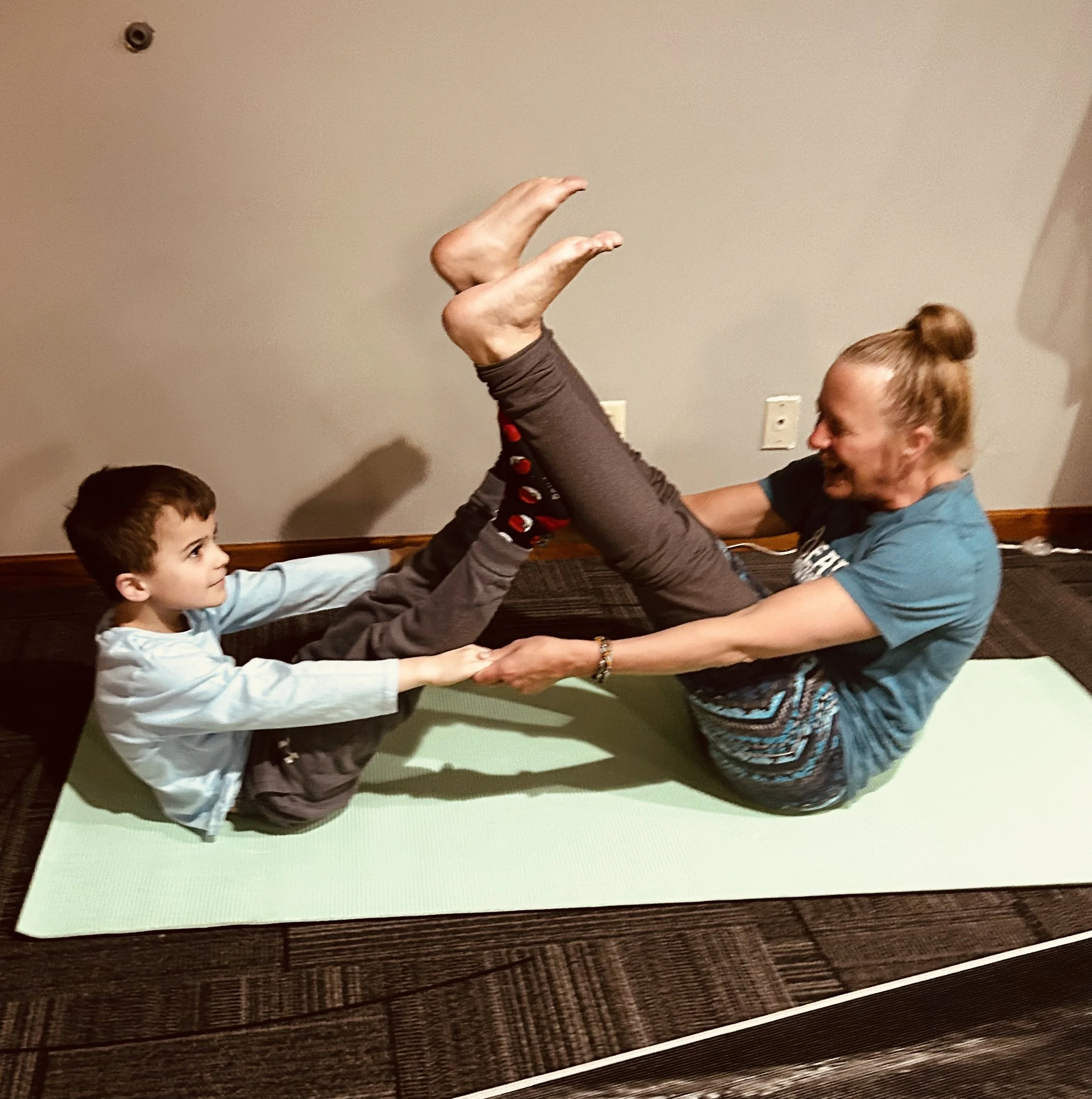 Two children and an adult engaging in a playful activity, holding the adult upside down by their legs while the children assist. The environment appears to be a room with a beige wall and electrical outlets visible.