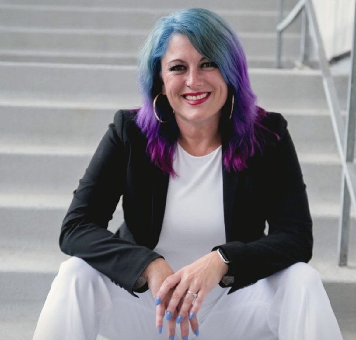 A woman with vibrant, multi-colored hair in shades of blue, purple, and pink, sitting on stairs, smiling, wearing a black blazer, white shirt, white pants, and hoop earrings.