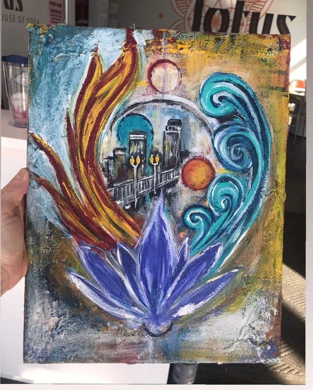 Colorful abstract painting of a cityscape with a bridge, a building labeled 'WOODMAN', and various swirling shapes, a large purple lotus flower at the bottom, and a vibrant background with blue, orange, yellow, and green hues.