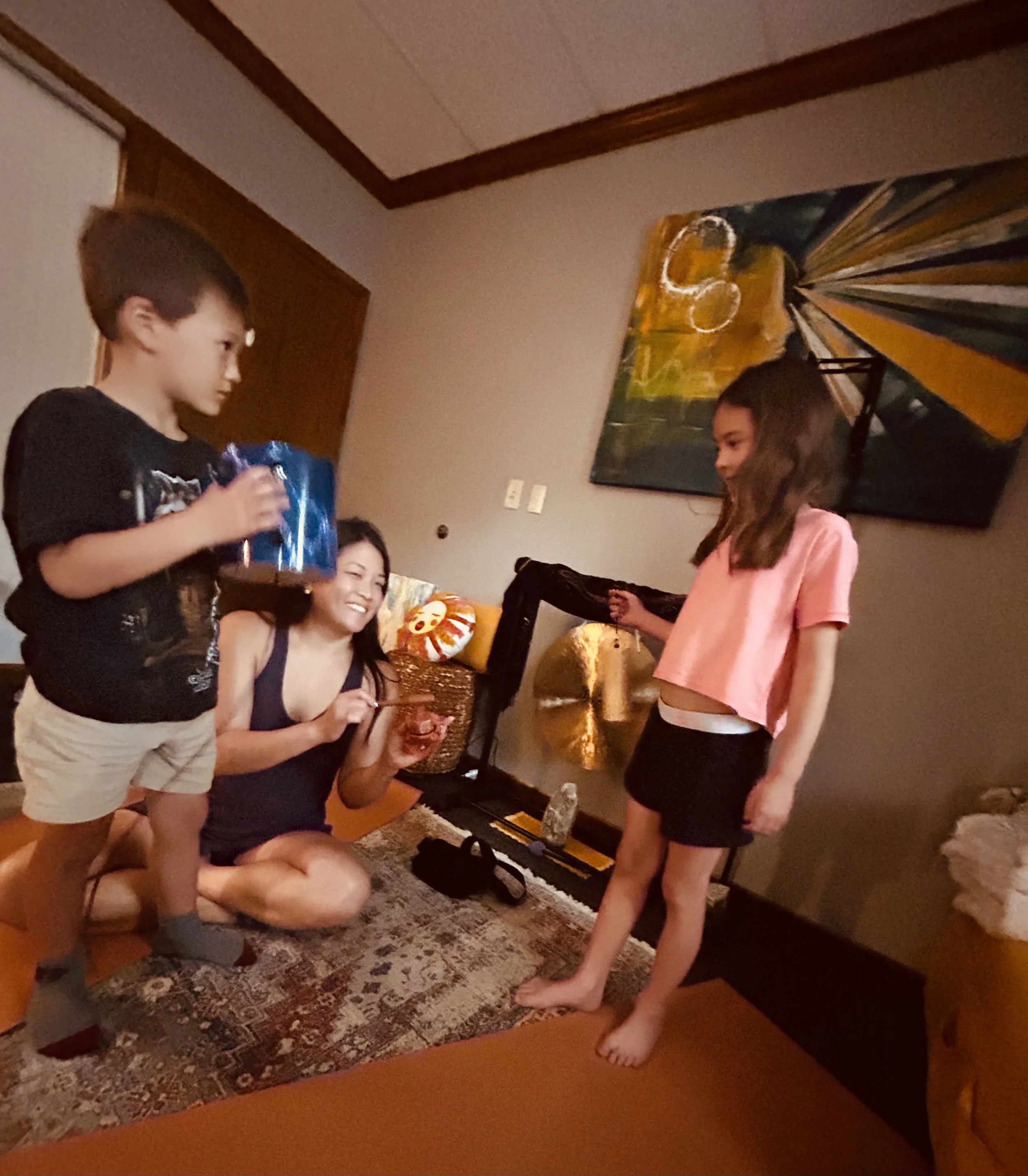 Children and adults gathering in a living room, with one girl in a pink shirt standing on the floor, others seated, and decorative pillows on a couch, in front of a large colorful abstract painting on the wall.