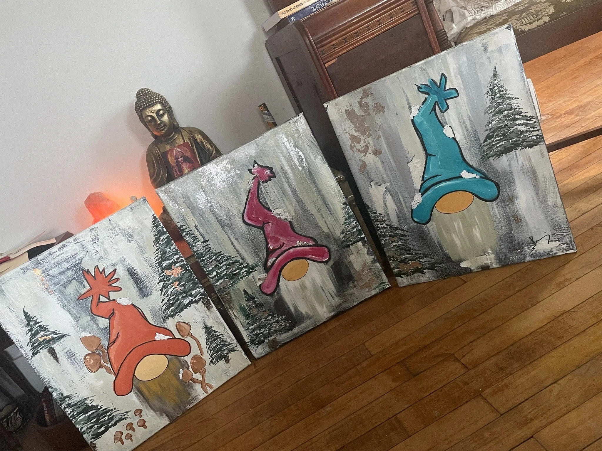 Four paintings of stylized dogs with snowy forest backgrounds and trees, with one painting showing a flame symbol, and another showing a peace sign, leaning against a wooden piece of furniture in a room.