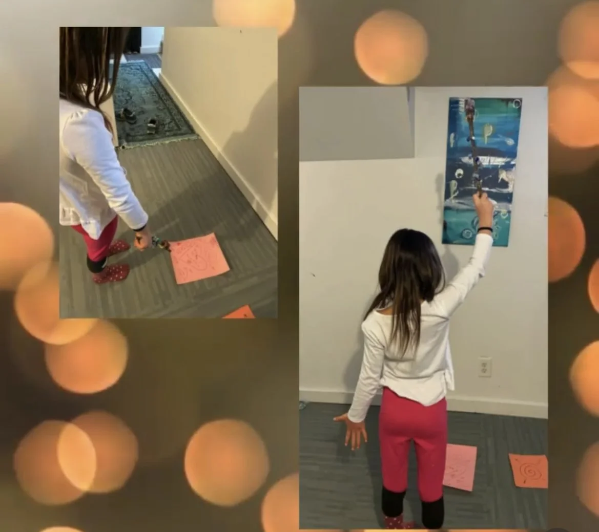 A girl drawing on a pink paper on the floor in front of a wall with artwork, with another girl standing and drawing on a canvas on the wall.