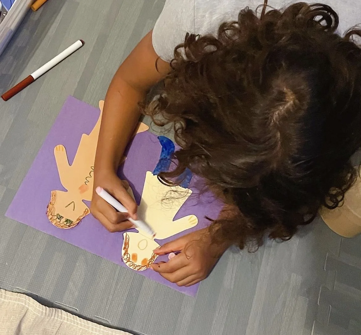 A person with curly brown hair creating a craft project on a purple poster board, drawing and coloring with markers. The project involves paper cutouts of hands with decorated palms.