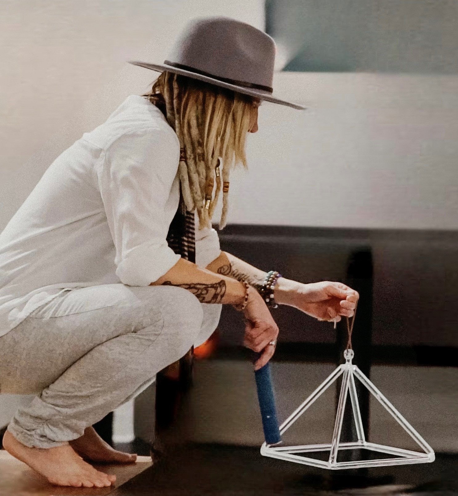 A woman with long dreadlocks wearing a wide-brimmed hat, white shirt, and beige pants, sits on a couch, looking down at a small skeleton figure hanging from a string attached to a tripod stand.