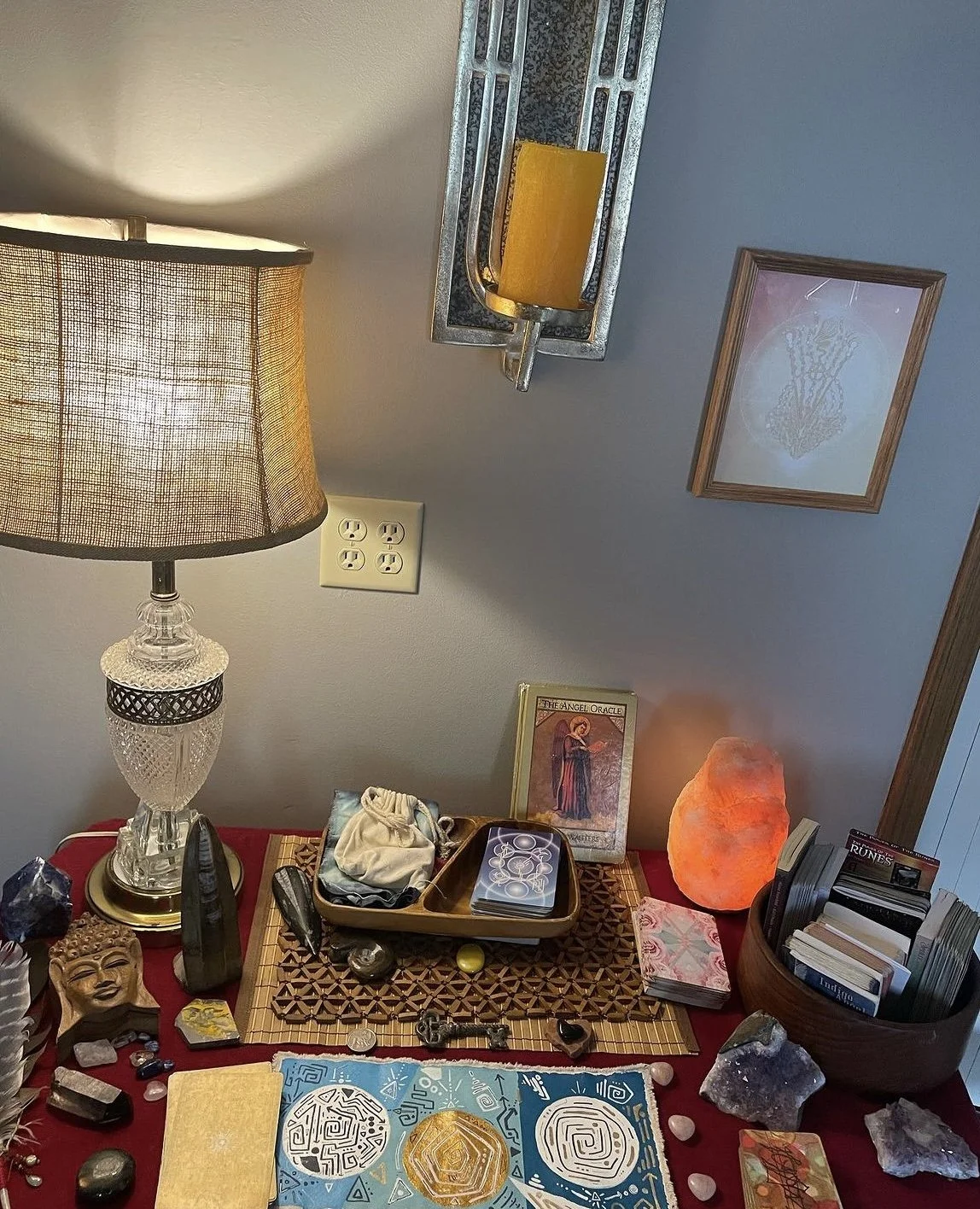A table with spiritual and decorative items, including a salt lamp, tarot cards, a Buddha head sculpture, crystals, and a framed picture. A lamp with a fabric shade is on the left side of the image, and a wall with framed artwork is visible in the ba