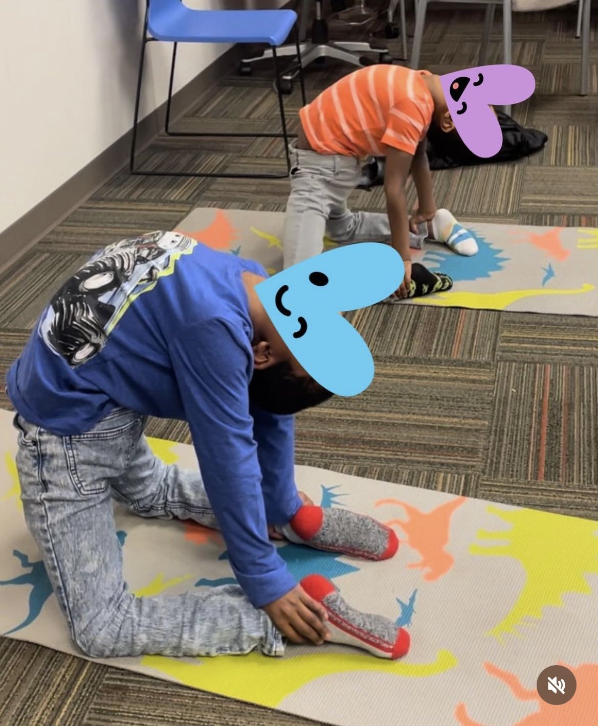Two children with cartoon heart-shaped emoji faces practicing stretches on colorful dinosaur-themed mats in a room with chairs and a carpeted floor.