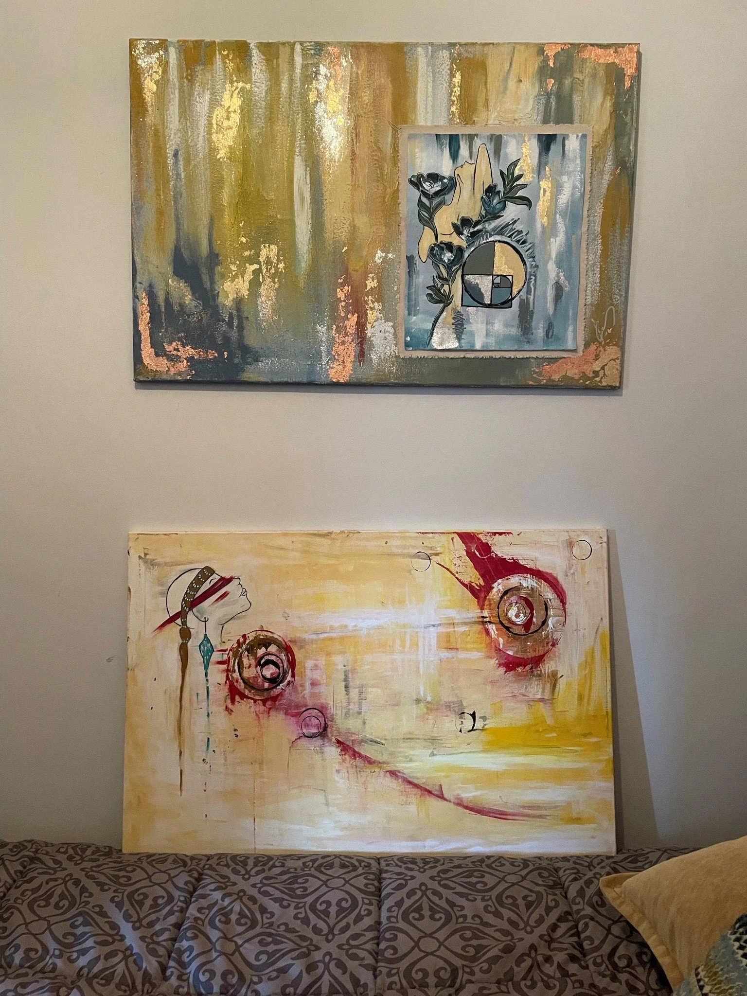 A large abstract painting hangs on a light-colored wall above a patterned couch and a pillow. The painting features a mix of gold, beige, gray, and pink tones with a central piece showing a floral design within a circle, resembling a Fibonacci spiral