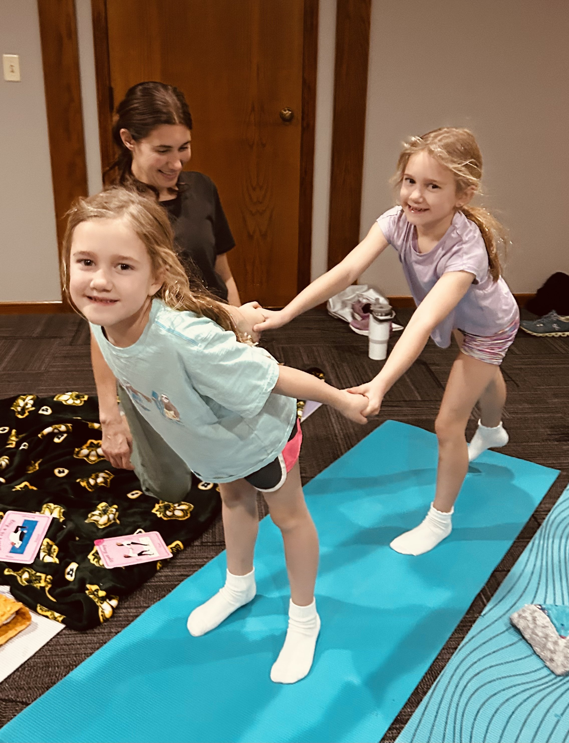 Two young girls are on a blue yoga mat holding hands in a playful pose, with an adult woman laughing in the background in a room with wood-paneled walls.