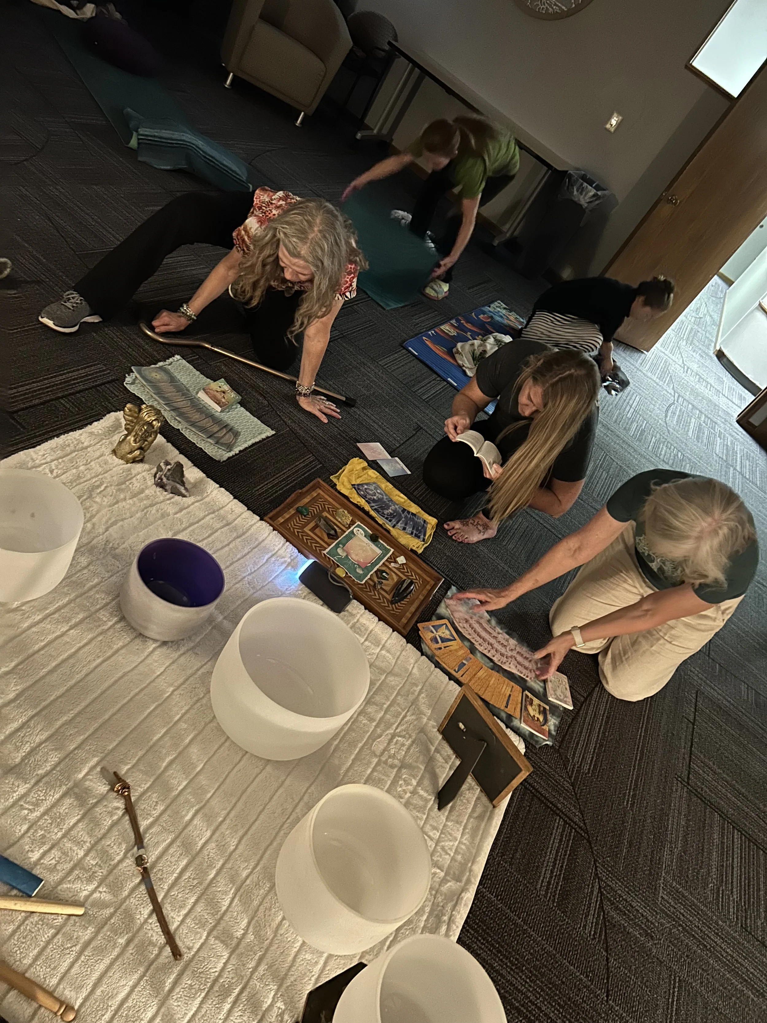 People sitting on the floor participating in a tarot card reading session, with a table in front holding tarot cards and decorative objects, in a room with dark carpet and beige carpeting, some chairs, and a door open to an outside area.