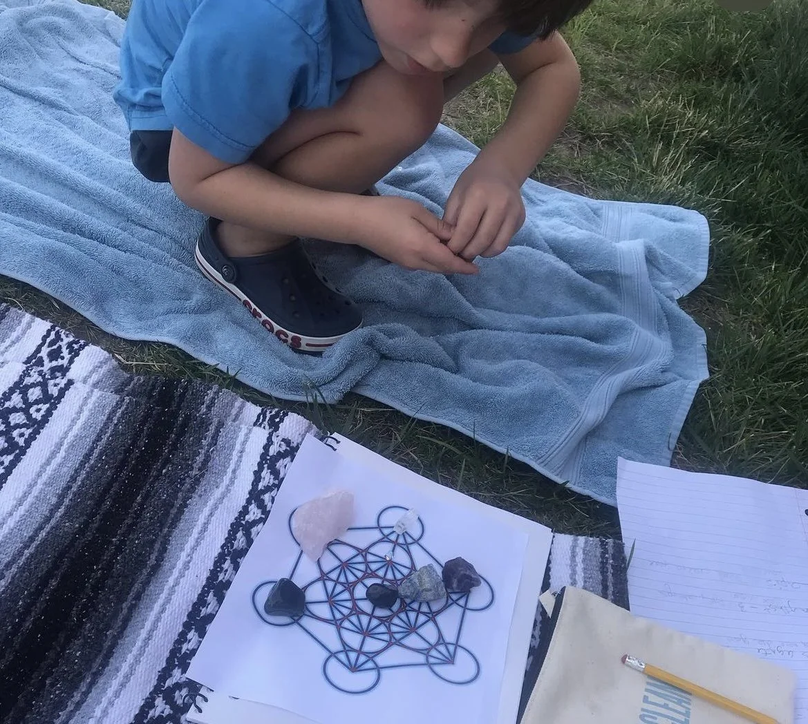 A young boy sitting cross-legged on a blue towel on the grass, with a rock-crystal grid laid out on paper in front of him, surrounded by various rocks, a pencil, and a notebook.