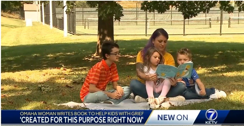 A woman with purple hair reading a children's book to three children sitting on a blanket under a tree in a park.