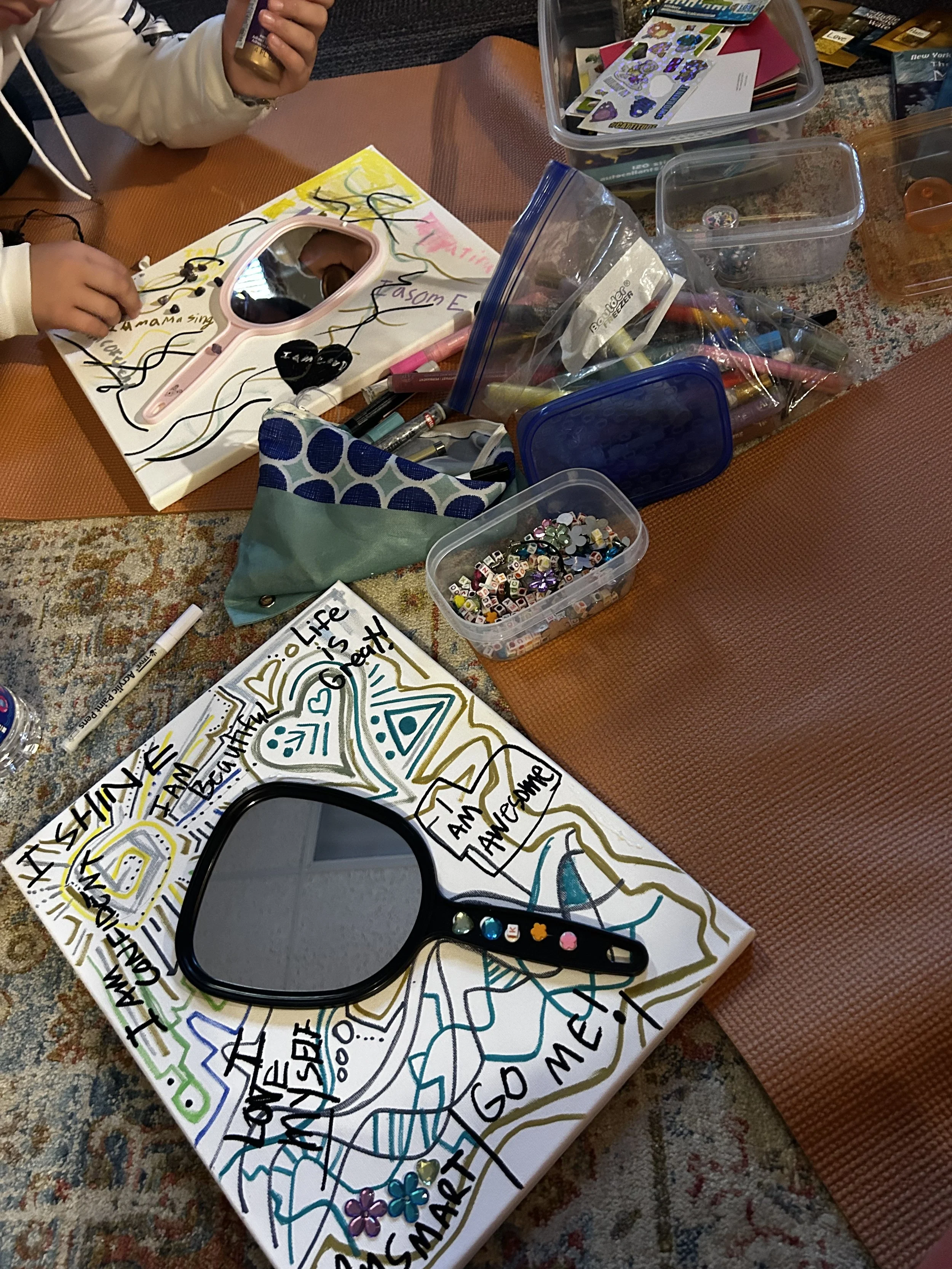 Colorful hand-drawn poster with words like 'fish', 'life', 'amazing', and 'greetings', surrounded by doodles, flowers, and patterns. Various art supplies, containers of markers, stickers, and decorated objects are on the table.