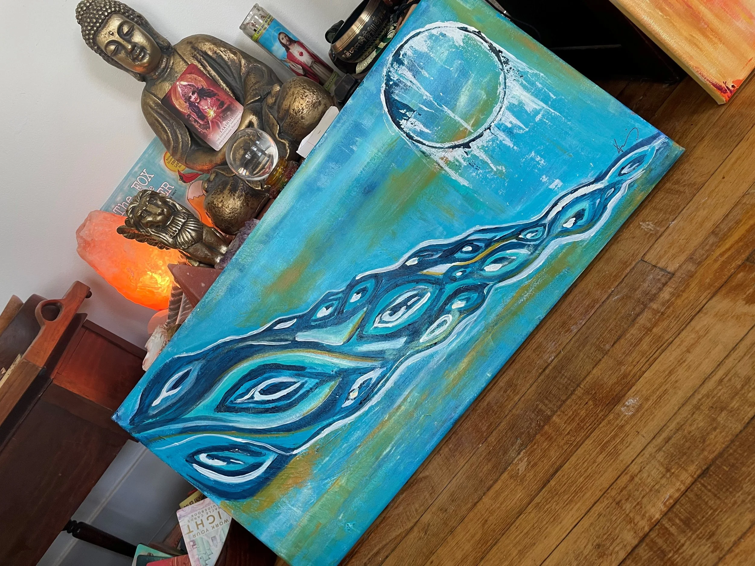 A wooden table with a painting of a rainbow-colored wave and a large circle in the sky. Behind the table, various statues, candles, and religious images are placed, including a Buddha statue and a smaller deity statue, along with salt lamps and spiri