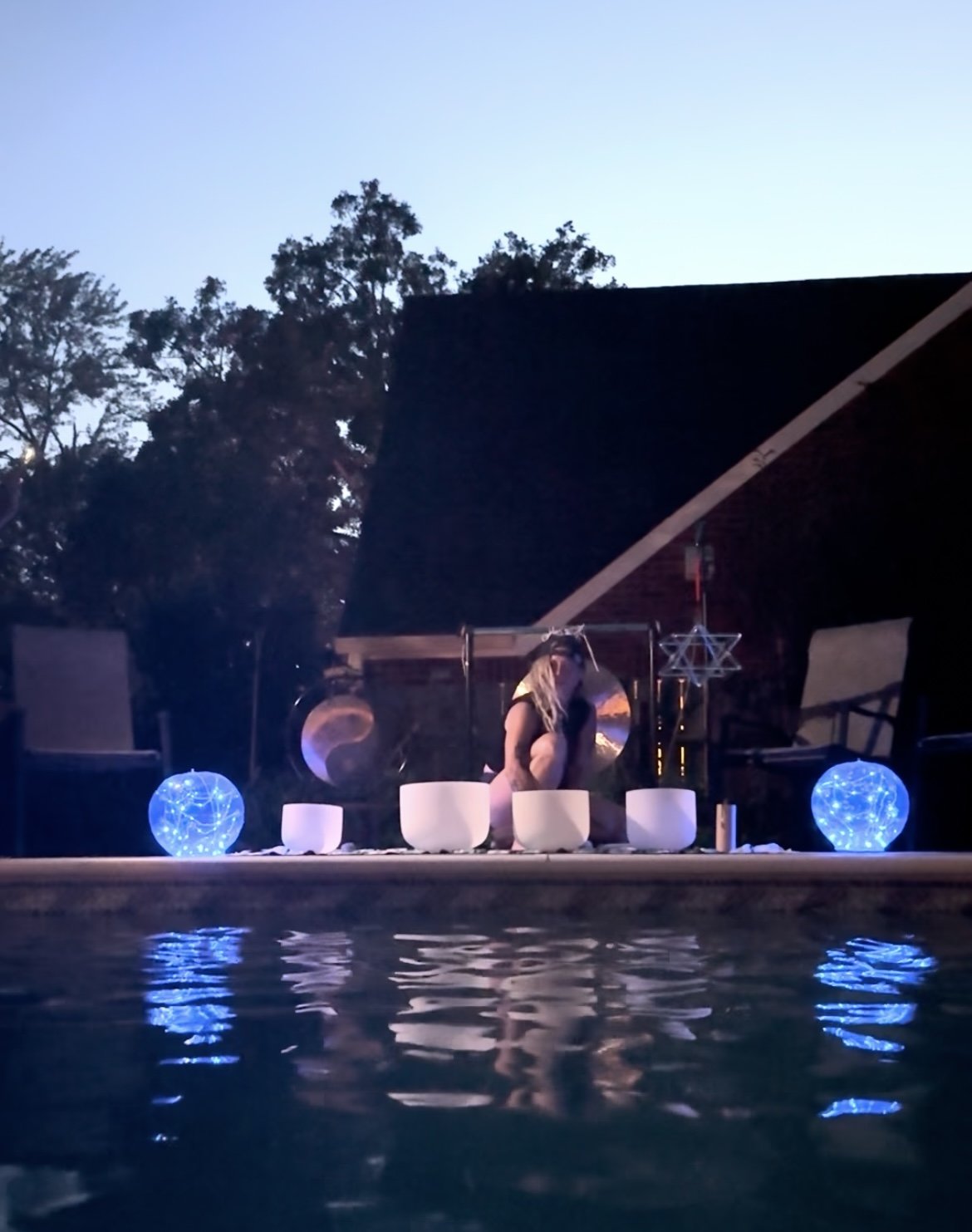 A woman with long blonde hair practicing Sound Healing on a platform by a pool during twilight. The platform is decorated with white singing bowls, illuminated blue orbs, and mystical stars and shapes, creating a serene and spiritual atmosphere.