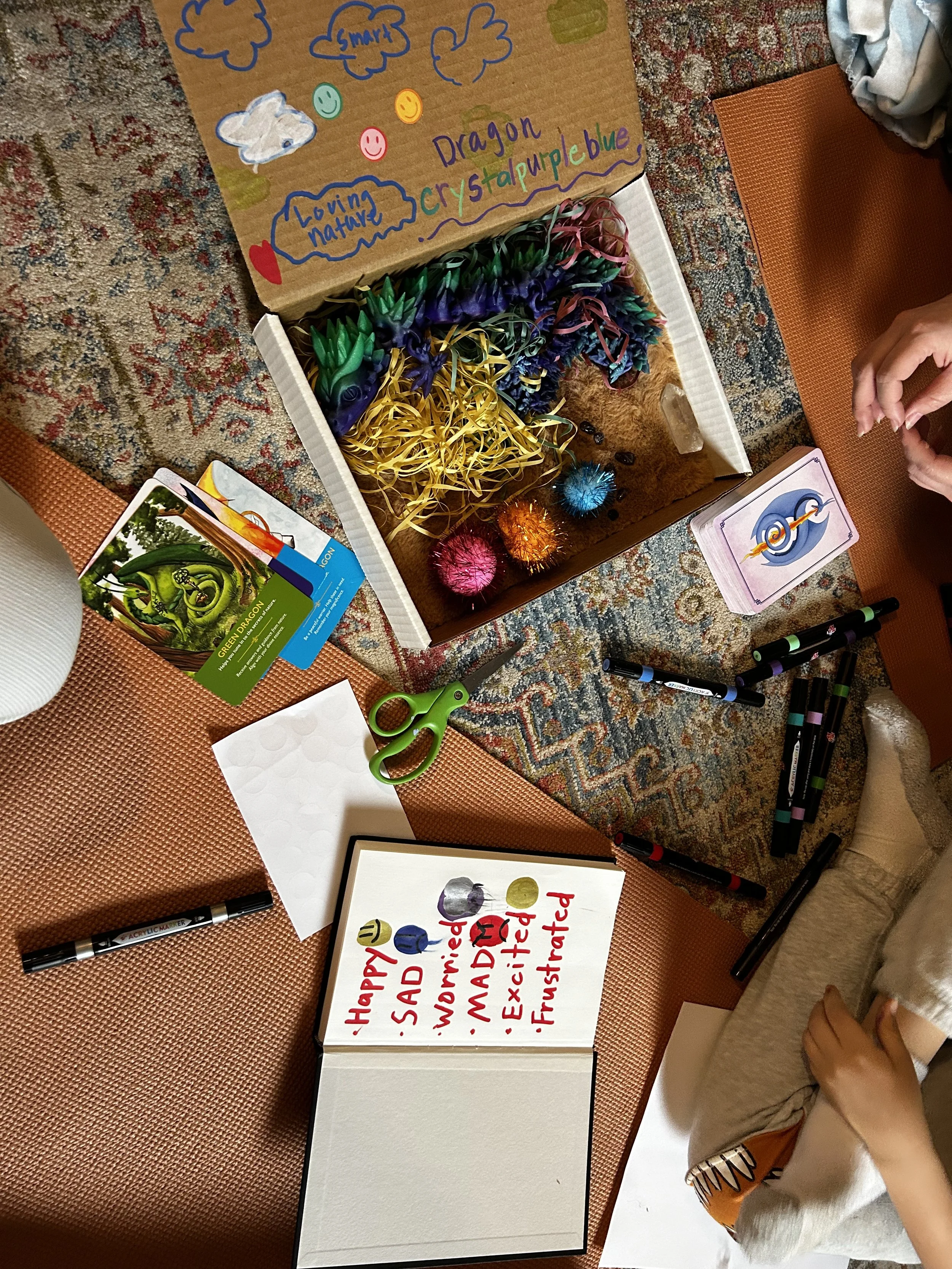 A classroom scene with a student’s hand writing on a whiteboard with colorful markers. The board has a list of words related to a holiday or celebration, including 'Happy,' 'Sad,' 'Worried,' 'Mad,' 'Excited,' 'Anxious,' and 'Frustrated'. Nearby are c