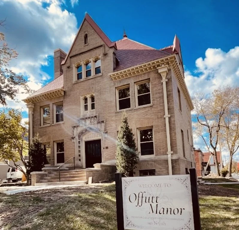 A large, three-story historic house made of light-colored brick with a steeply pitched roof, decorative trim, and multiple windows. A sign in front reads "Welcome to the Offutt Manor." The scene is set outdoors with a partly cloudy sky and trees.