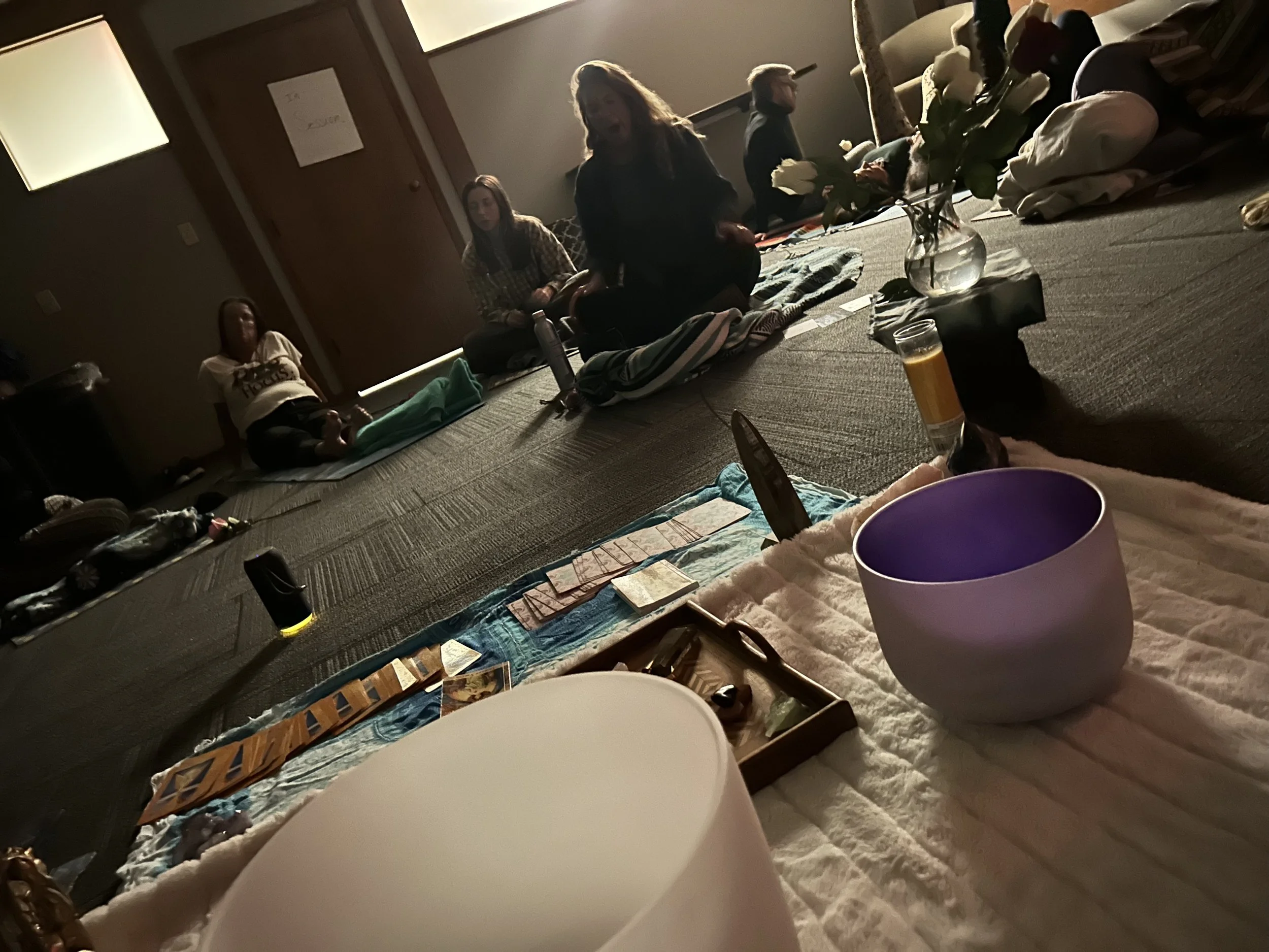 Group sound bath meditation. People sitting on the floor in a dimly lit room, gathered around cards, flowers, and various personal items.