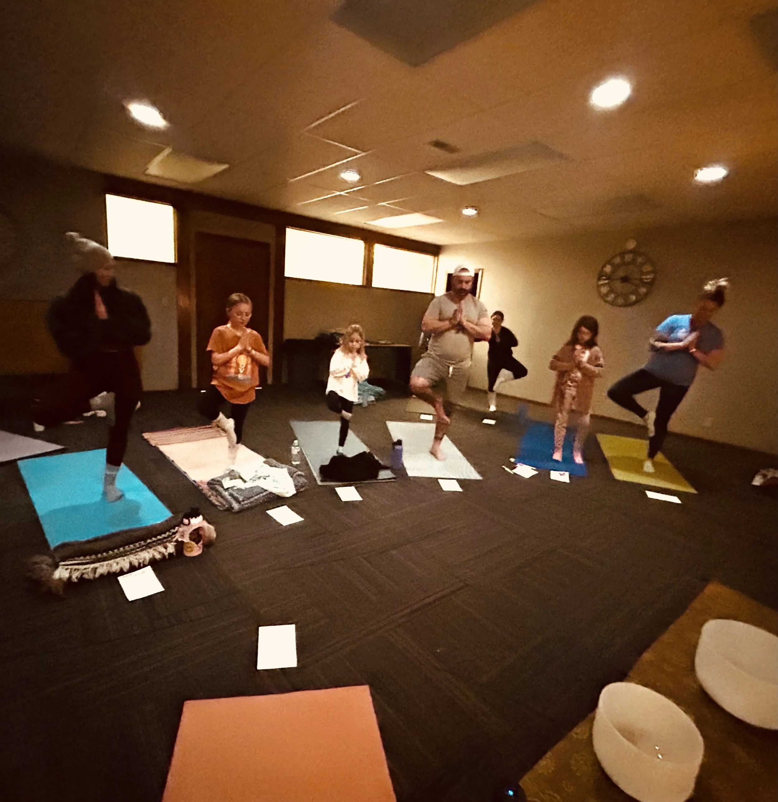 People practicing yoga in a dimly lit room with colorful yoga mats and a clock on the wall.