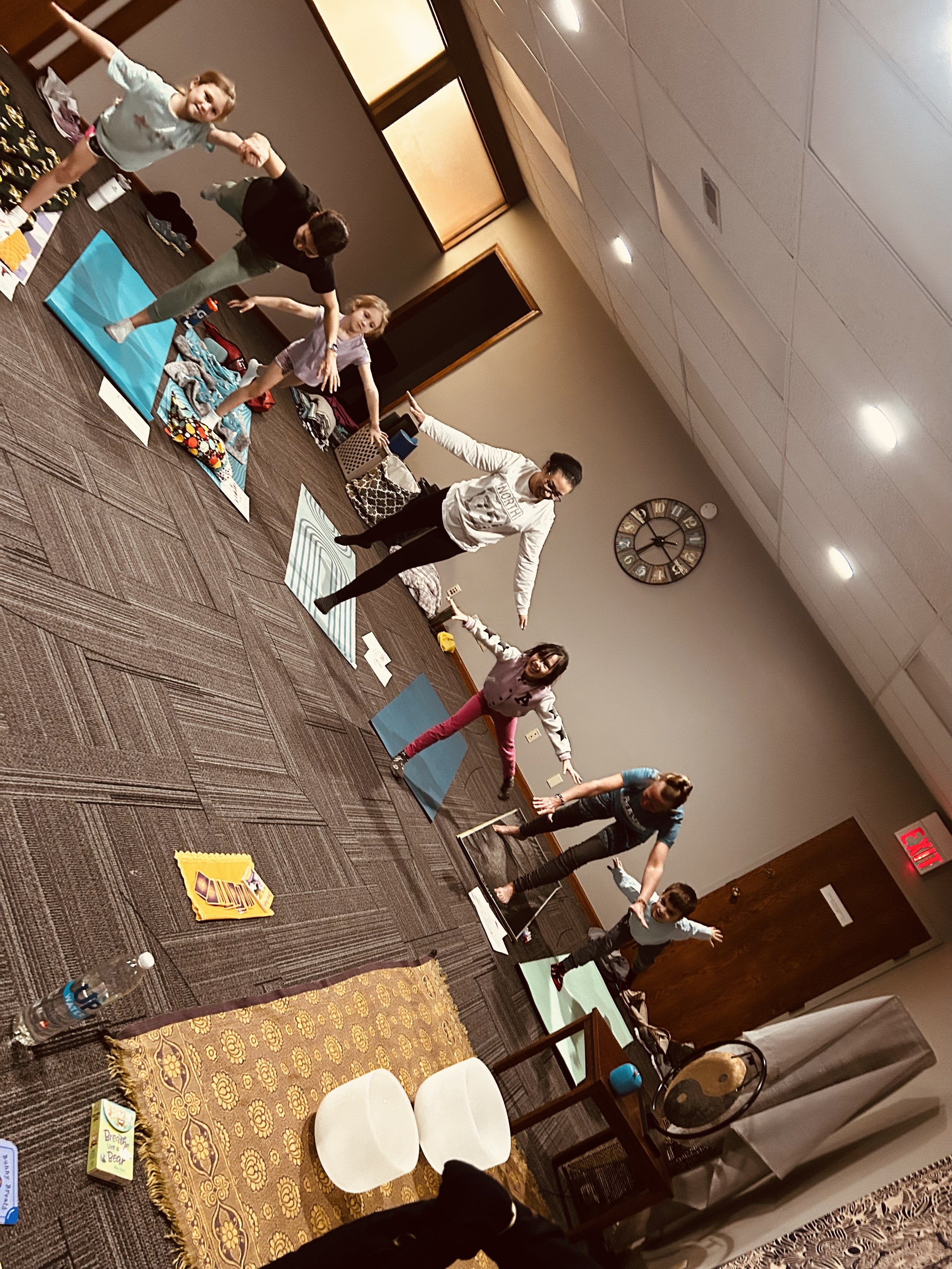 Group of children and adults participating in a stretching or yoga activity in a room with tables and chairs, mats, and various items on the table, including water bottles and games.