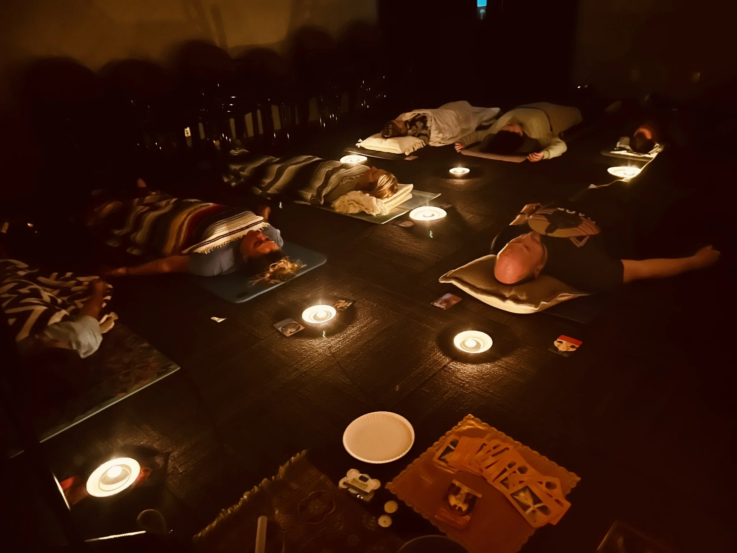 People lying on the floor with pillows and blankets in a dark room illuminated by small candles, possibly engaging in a group meditation or relaxation session.