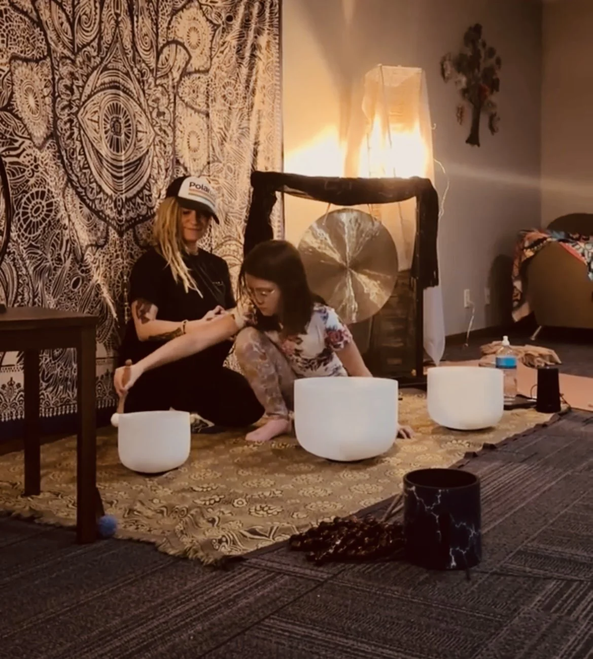 Two women sitting on the floor with singing bowls in front of them, one woman wearing a hat, in a room with bohemian decor including tapestries and a large gong.