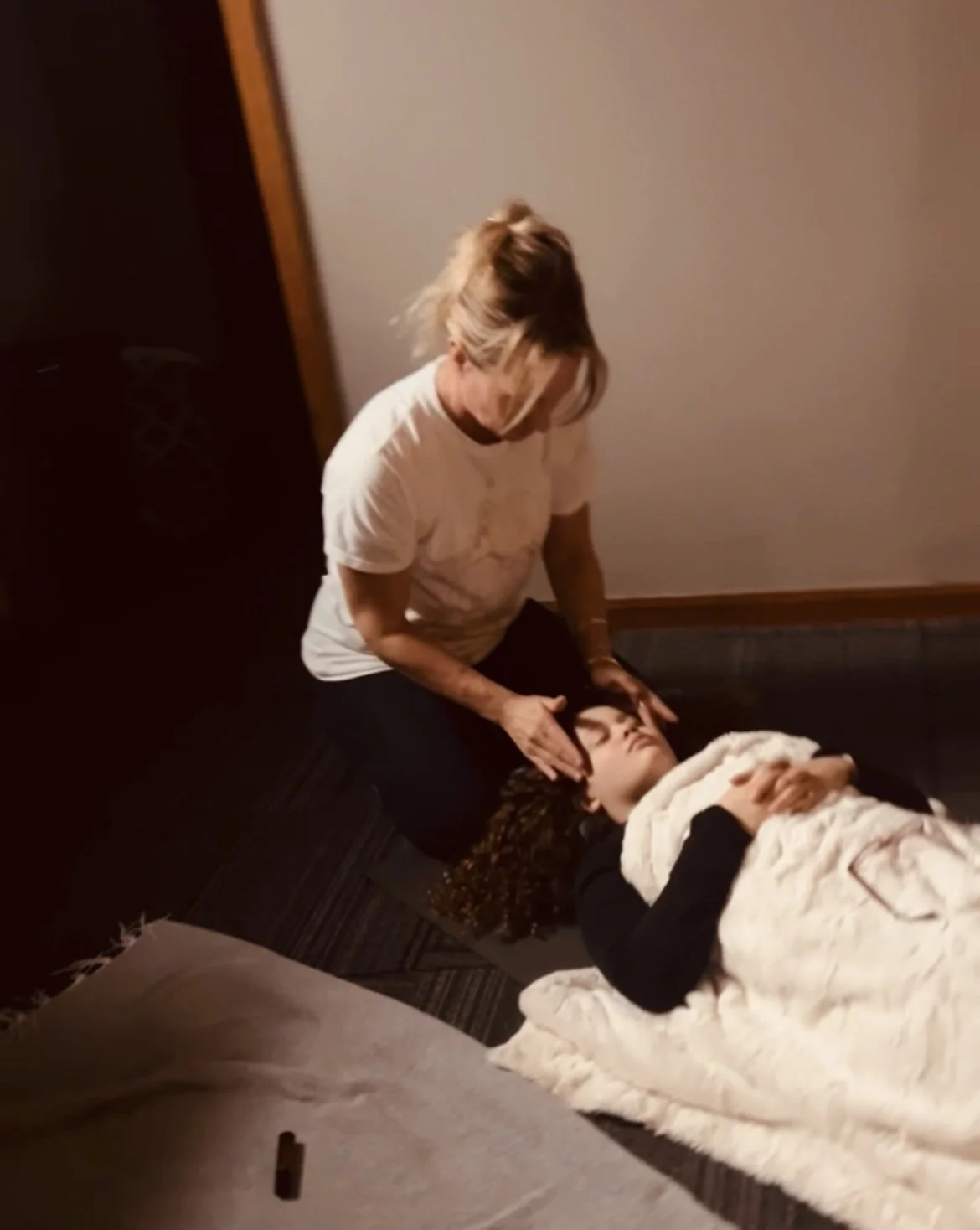A woman receiving a head massage from another woman lying on a bed with a white blanket, in a dimly lit room.