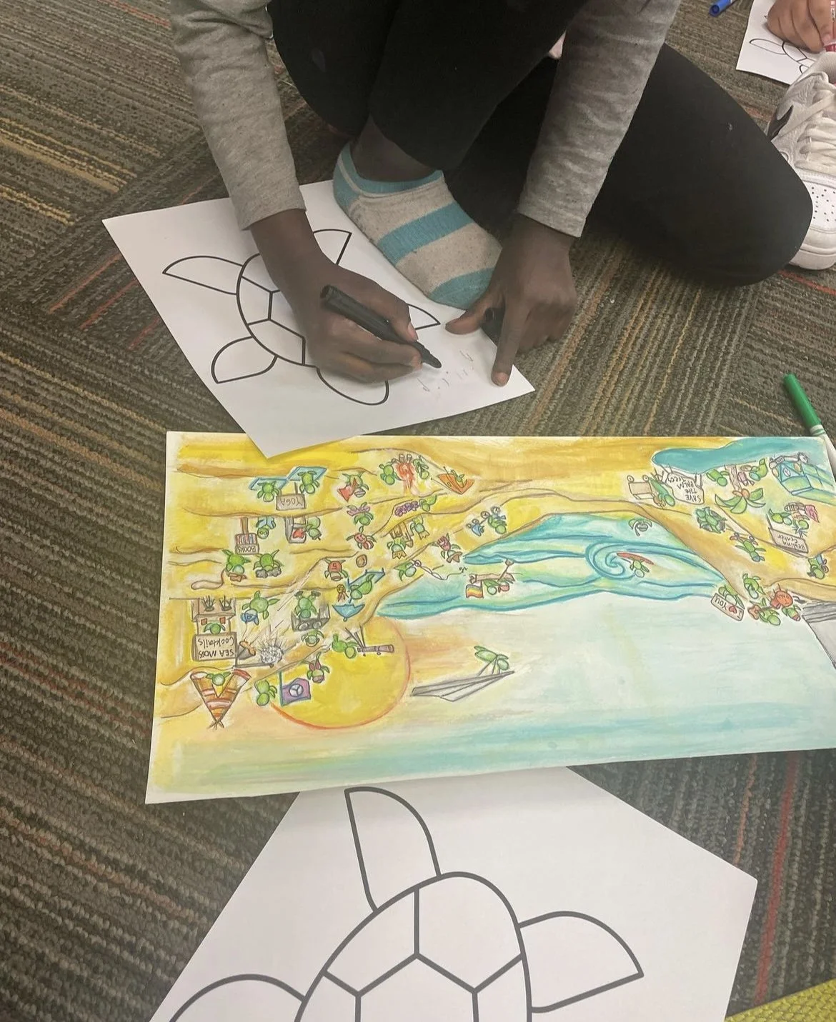 Child coloring a beach scene with a yellow sun, blue water, and trees, while sitting on a carpeted floor with drawings and supplies around.
