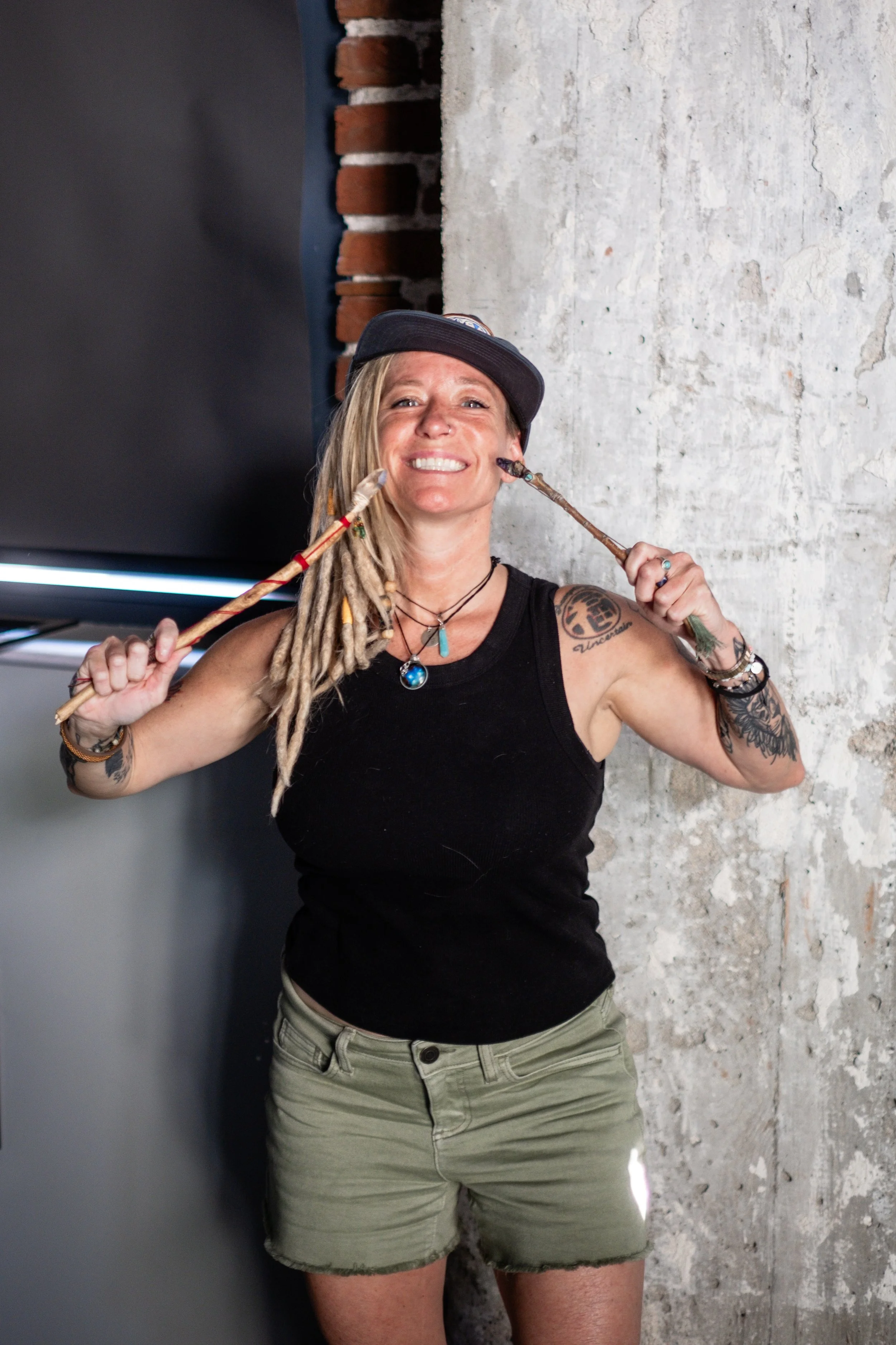 A woman with dreadlocks wearing a black tank top, khaki shorts, a baseball cap, and jewelry, standing against a concrete wall and holding sticks near her face with a big smile.