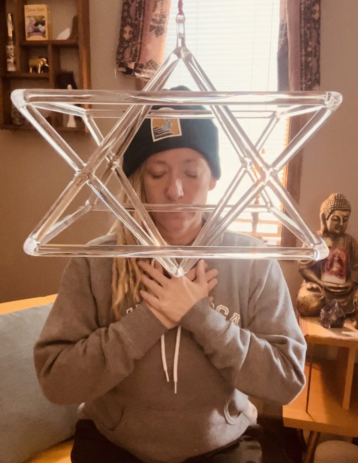 A person with long blonde hair, wearing a black beanie and a gray hoodie, is sitting with their eyes closed and hands crossed over their chest, behind a clear geometric plastic star sculpture. There is a Buddha statue and a wooden shelf with smaller decorative items in the background.