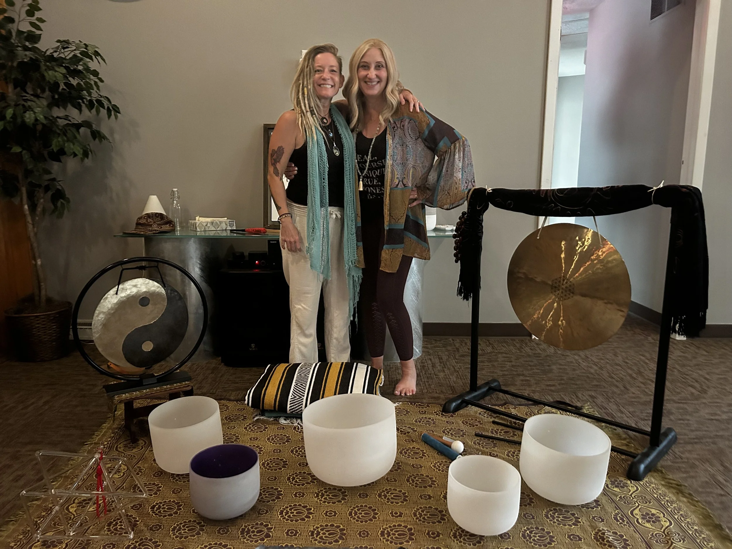 Two women stand together in a room decorated with singing bowls, a gong, and a black cloth. They are smiling, with one woman having dreadlocks and the other with blonde hair. There are various objects on the table behind them, including a small lamp 