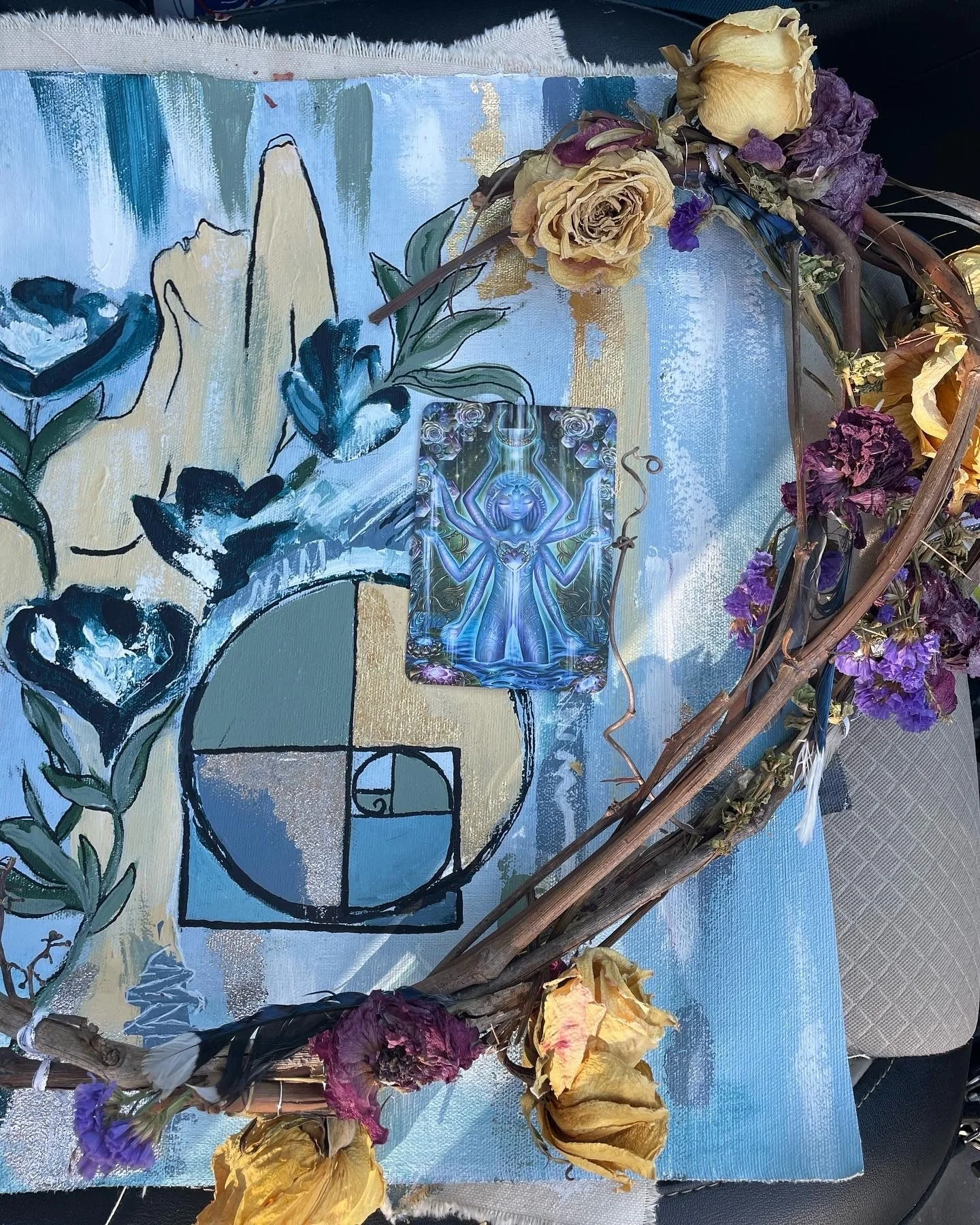 A painted canvas with a geometric and abstract floral design, decorated with a wreath of dried flowers and leaves, and a tarot card featuring a mystical figure with multiple arms.