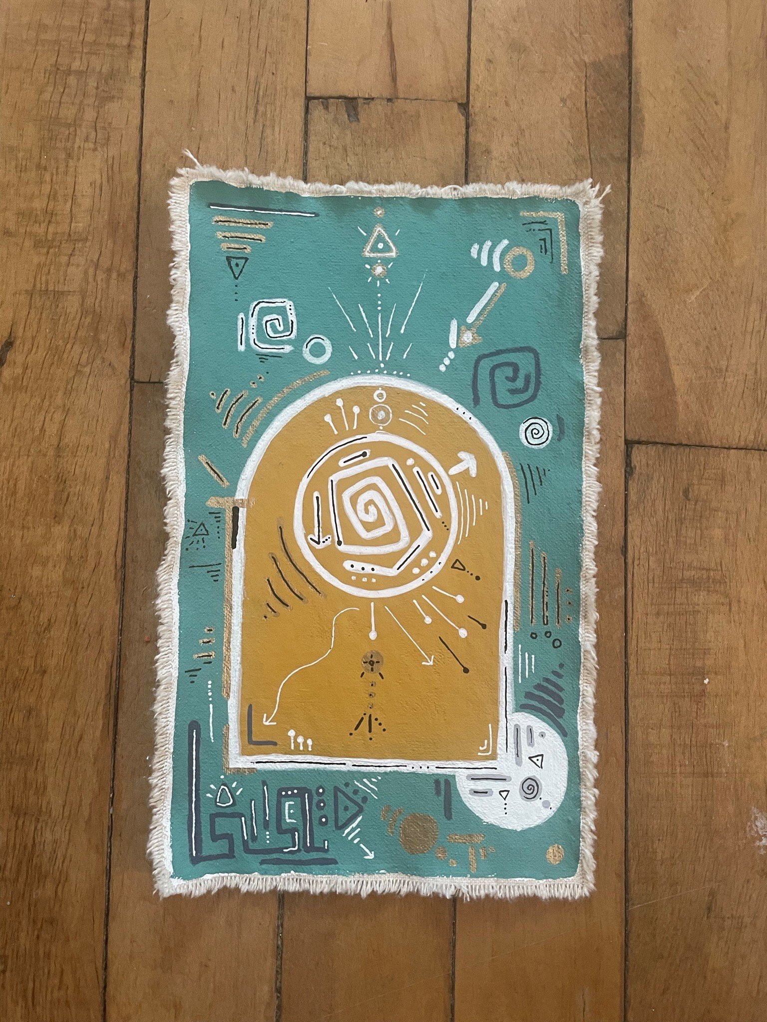 A decorative rectangular rug with abstract patterns in white, gold, and black on a green background, fringed edges, placed on a wooden floor.