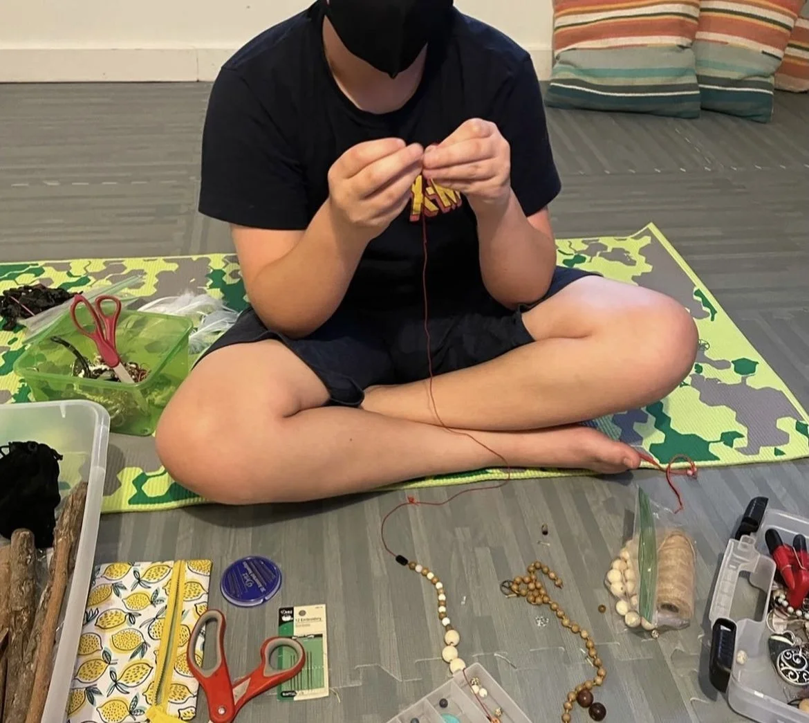 A person sitting cross-legged on a camouflage-patterned yoga mat, making jewelry with various beads and tools organized around them on the floor.