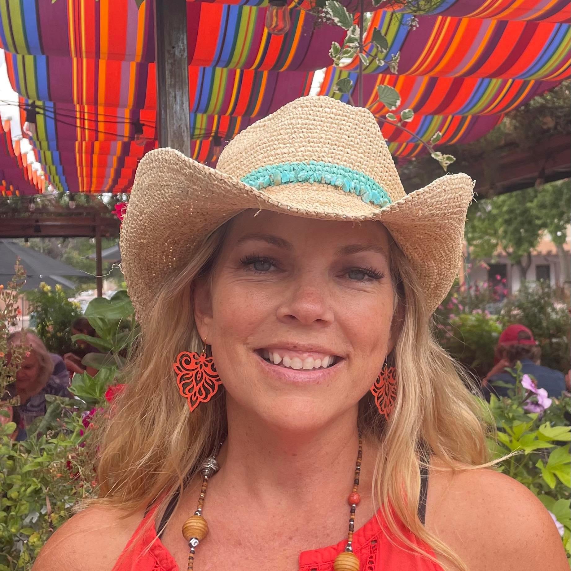 A woman with blonde hair, blue eyes, and a bright smile, wearing a woven cowboy hat with a blue decorative band. She is also wearing large orange butterfly earrings, a beaded necklace, and a red sleeveless top. The background features colorful striped fabric or umbrellas and greenery with other people visible.