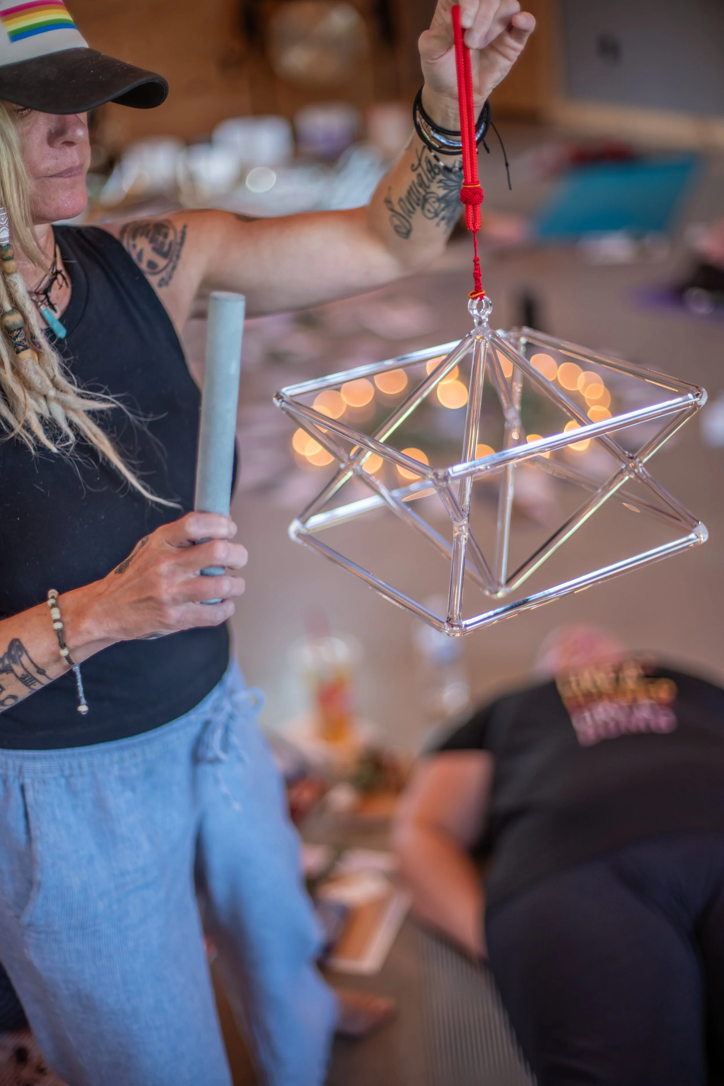A woman with tattoos on her arm is holding a red rope and a transparent star-shaped lantern with lights inside, in a room with wood walls and other people in the background.