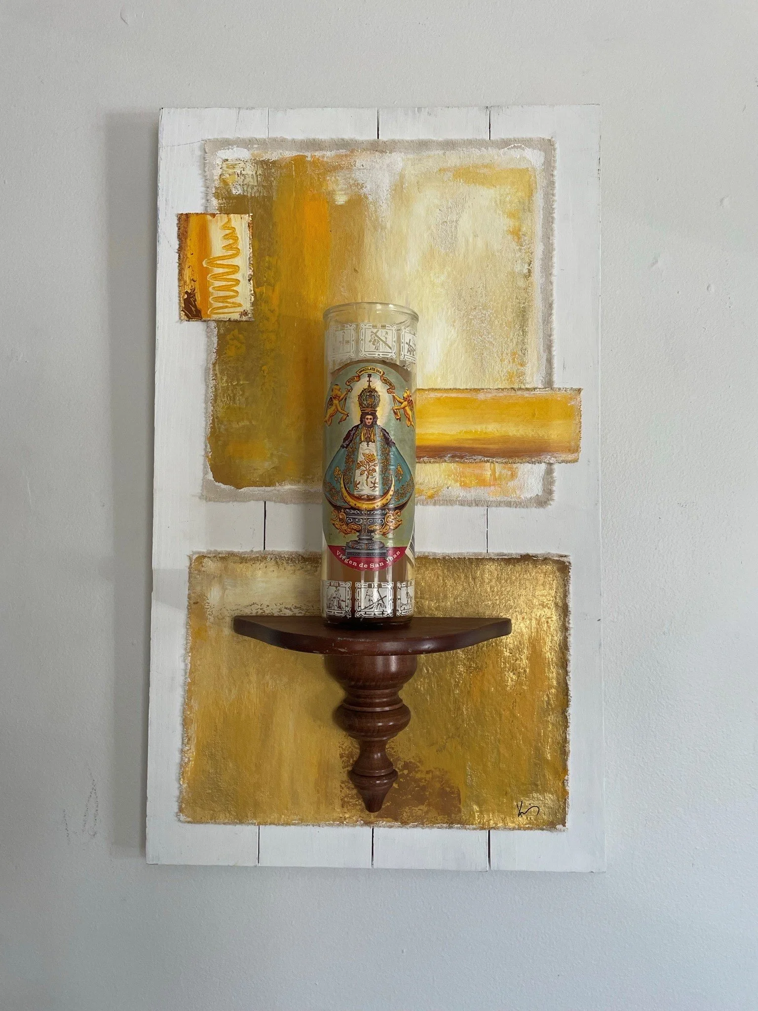 Decorative religious candle holder with an image of the Virgin of San Juan on a carved wooden base, mounted on an abstract yellow and white textured painting.